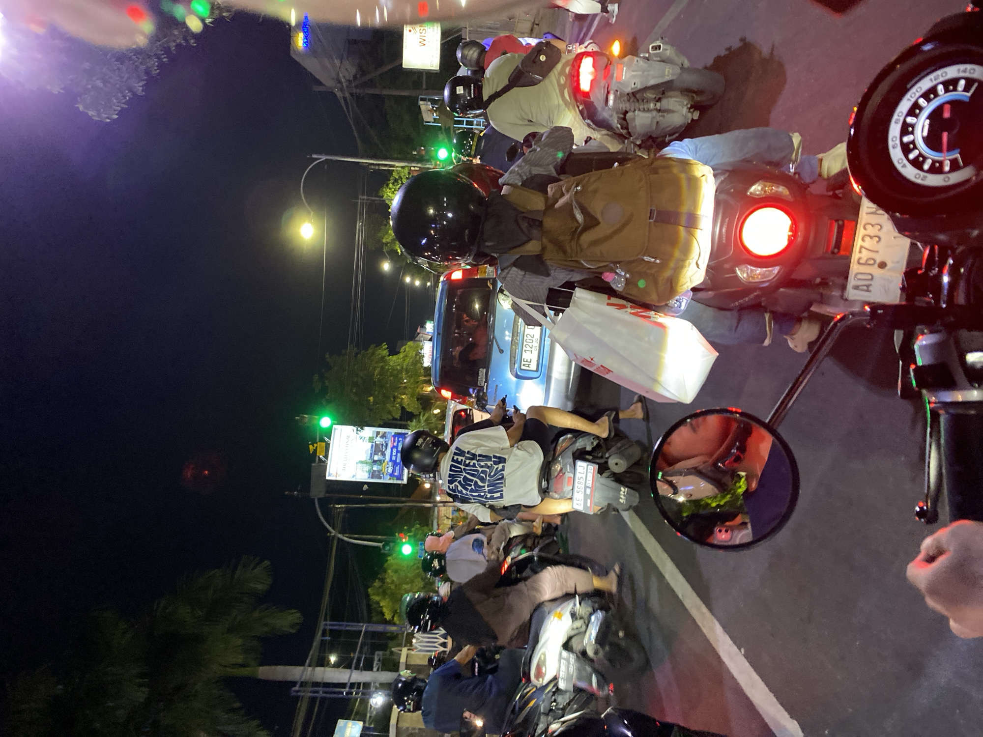The image captures a vibrant night scene on a busy street in Madiun, Indonesia, viewed from the perspective of a person on a motorbike, likely waiting in traffic. Numerous motorbikes, mostly scooters, are stopped in rows, along with a blue car further down the road. All motorbike riders are wearing helmets, and several carry backpacks or shopping bags.
The setting is an urban environment illuminated by various light sources: vehicle headlights and taillights, bright streetlights, and green traffic signals, indicating the time of day is night. The sky is dark, and the weather appears clear. In the immediate foreground, the dashboard and speedometer of the motorbike from which the photo is taken are visible, showing numbers up to "140". A rearview mirror on the right reflects a blurry image.
Visible text includes several Indonesian license plates: "AE 5685 LJ", "AE 1202 D", and "AD 6733 N". A commercial sign on the left displays "WISMA" and partially "THE LUFVVFINAL". The back of one person's white t-shirt reads "ENDEV EMIE". The scene portrays the common sight of heavy motorbike traffic in Southeast Asian cities.