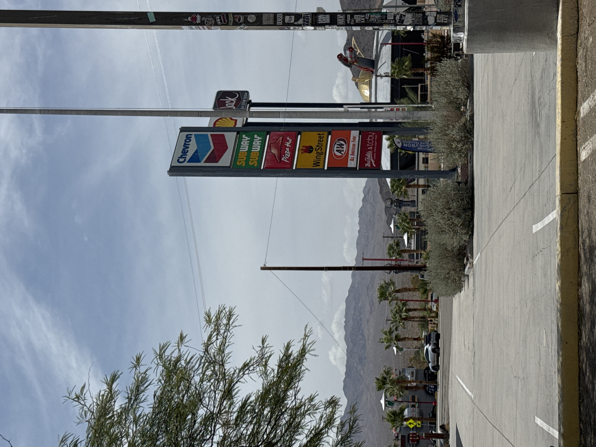 The image depicts a roadside service area in Baker, California, United States. A prominent signpost displays logos for Chevron, Subway, Pizza Hut, Wingstop, A&W, and Marie Callender's & Toby's. Adjacent to this sign, a Jack in the Box sign is visible. Further to the right, a large pole is covered in numerous stickers. In the background, a cluster of palm trees and desert vegetation is set against a backdrop of arid mountains. A white SUV is parked in the foreground on the left, and another vehicle is visible further back. The sky is overcast with scattered clouds. The overall scene suggests a typical highway rest stop in a desert environment.