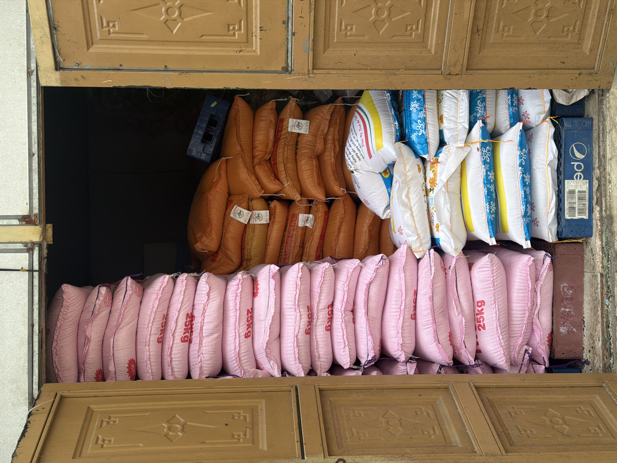 A street-level view in Đà Nẵng, Vietnam, shows a doorway filled with stacks of rice sacks. The door is a two-panel design with decorative carvings and a weathered appearance, suggesting age. The rice sacks are the main focus, arranged in three distinct layers. The bottom layer consists of pink sacks, each labeled "25kg." The middle layer shows orange sacks, while the top layer features white and blue patterned sacks. Blue plastic crates labeled "Pepsi" are used as support for the upper layers. The overall scene suggests a small business or storage location.
