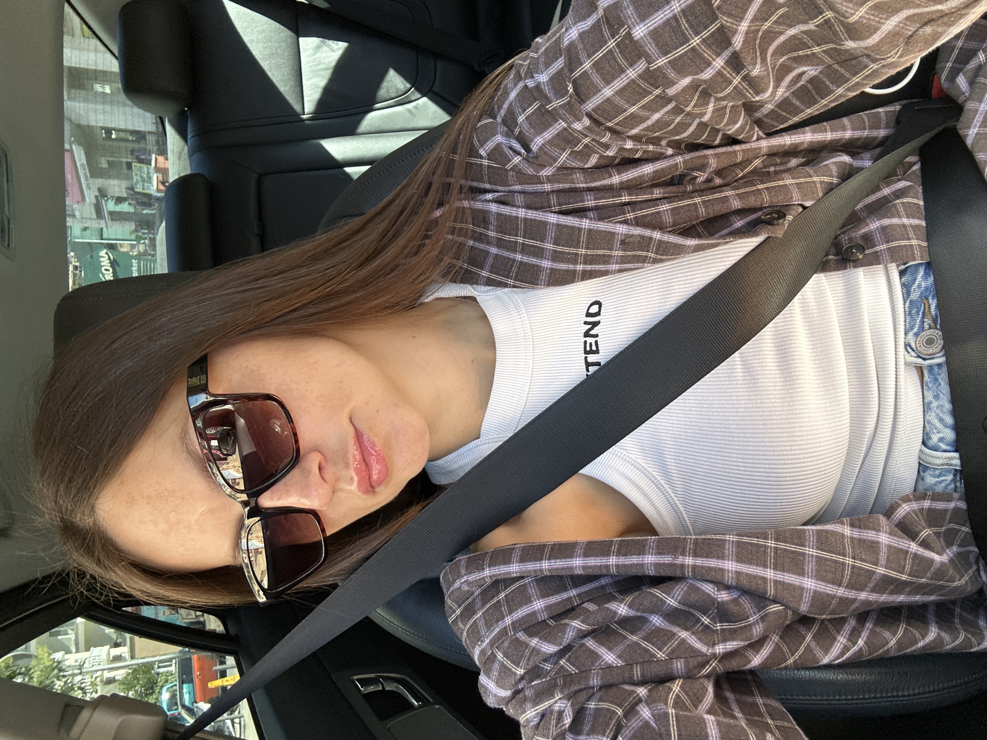 In Donji Pobori, Montenegro, a young woman with long brown hair, wearing large sunglasses, a white ribbed tank top with "TEND" printed on it, a plaid shirt unbuttoned, and blue jeans, is seated in the passenger seat of a car. She is wearing a black seatbelt across her torso. Her reflection is visible in her sunglasses. The interior of the car, including the black leather seats and the seatbelt, is clearly depicted. Outside the car window, buildings and trees are visible, indicating an urban or suburban environment. The sunlight casts shadows on the car's interior.
