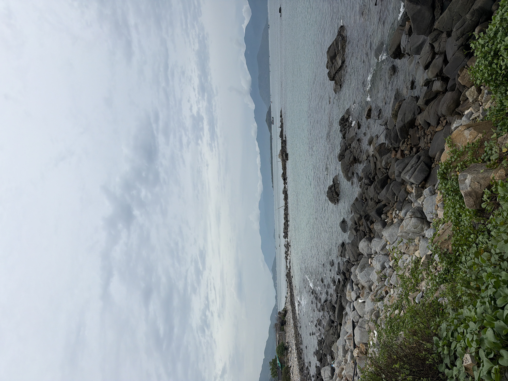 This image depicts a serene coastal landscape, likely in Vĩnh Lương, Vietnam, on an overcast day. The scene is divided horizontally into distinct layers, showcasing sky, sea, mountains, and a rocky shoreline.
The top portion of the frame is dominated by a wide, cloudy sky. The clouds are a mix of soft white and light grey, indicating an overcast condition without direct sunlight, creating diffused, muted lighting across the entire scene.
Below the sky, the calm, grey-blue sea stretches into the middle distance. Its surface shows subtle ripples near the shore, reflecting the subdued light from above. Along the horizon, a range of mountains rises, appearing hazy and pale blue due to atmospheric perspective, partially blending with the clouds and giving a sense of vastness. Very faint, indistinct shapes, possibly distant boats or buoys, can be seen on the water near the base of the mountains.
The foreground and right side of the image are characterized by a rugged, rocky coastline. Numerous dark grey boulders and smaller stones of various sizes form the shore, some partially submerged in the shallow waters where gentle waves break, creating sparse white foam. Interspersed among these rocks, particularly on the far right, are patches of vibrant green foliage and bushy coastal vegetation, providing a contrasting splash of color to the otherwise cool palette.
There are no people or discernible man-made objects present in the scene, which emphasizes its natural and untouched character. The time of day appears to be daytime, given the light, but the overcast conditions prevent a more precise determination. The overall atmosphere is one of tranquility and natural beauty. No text is visible in the image.