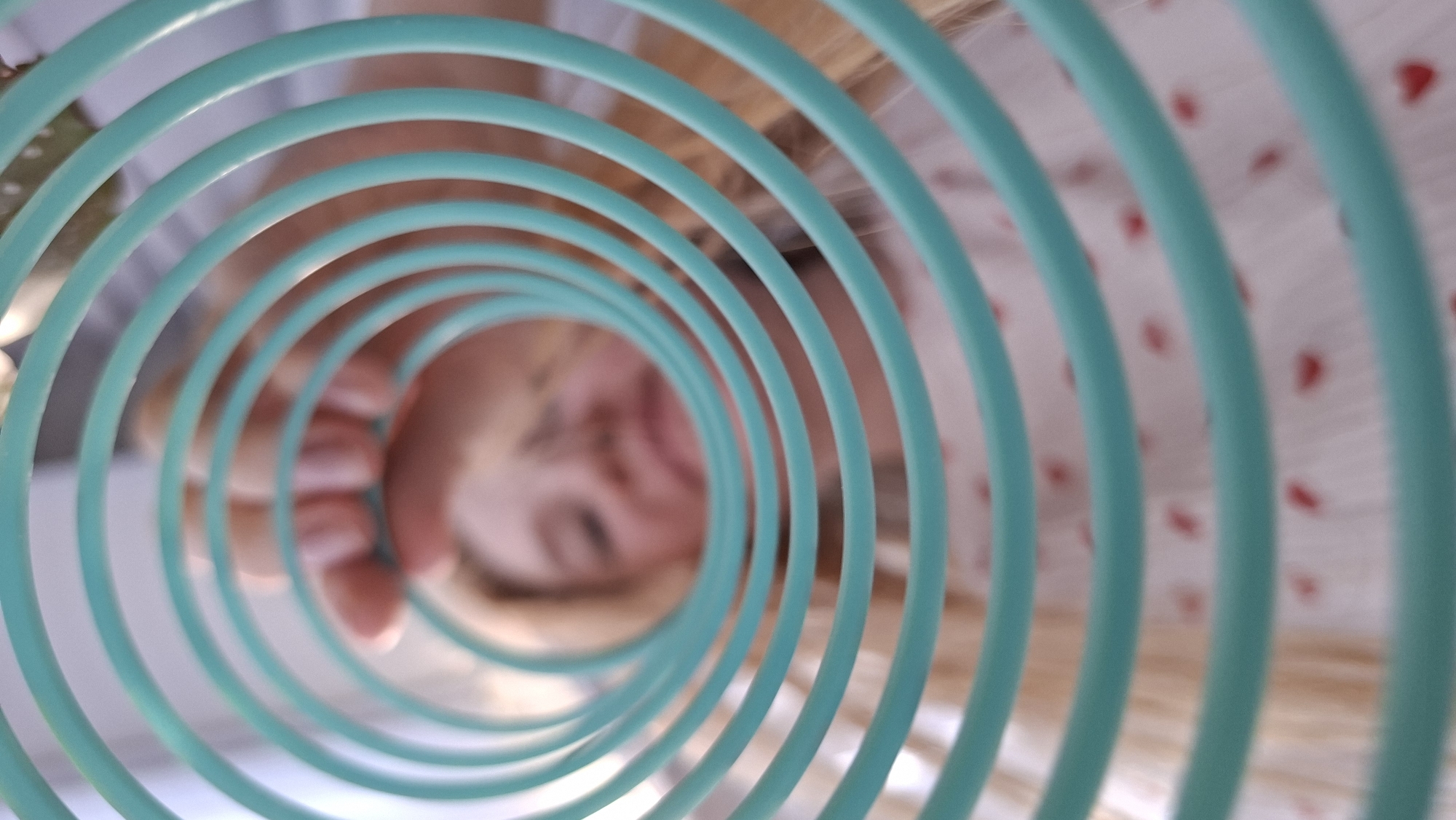 A low-angle shot looking up through a turquoise, coiled, plastic toy. A child with blonde hair and blurred facial features is visible through the center opening of the coil. The child is wearing a white garment with small red heart-shaped spots. The location is unidentifiable as Chaikovsky, Russia.
