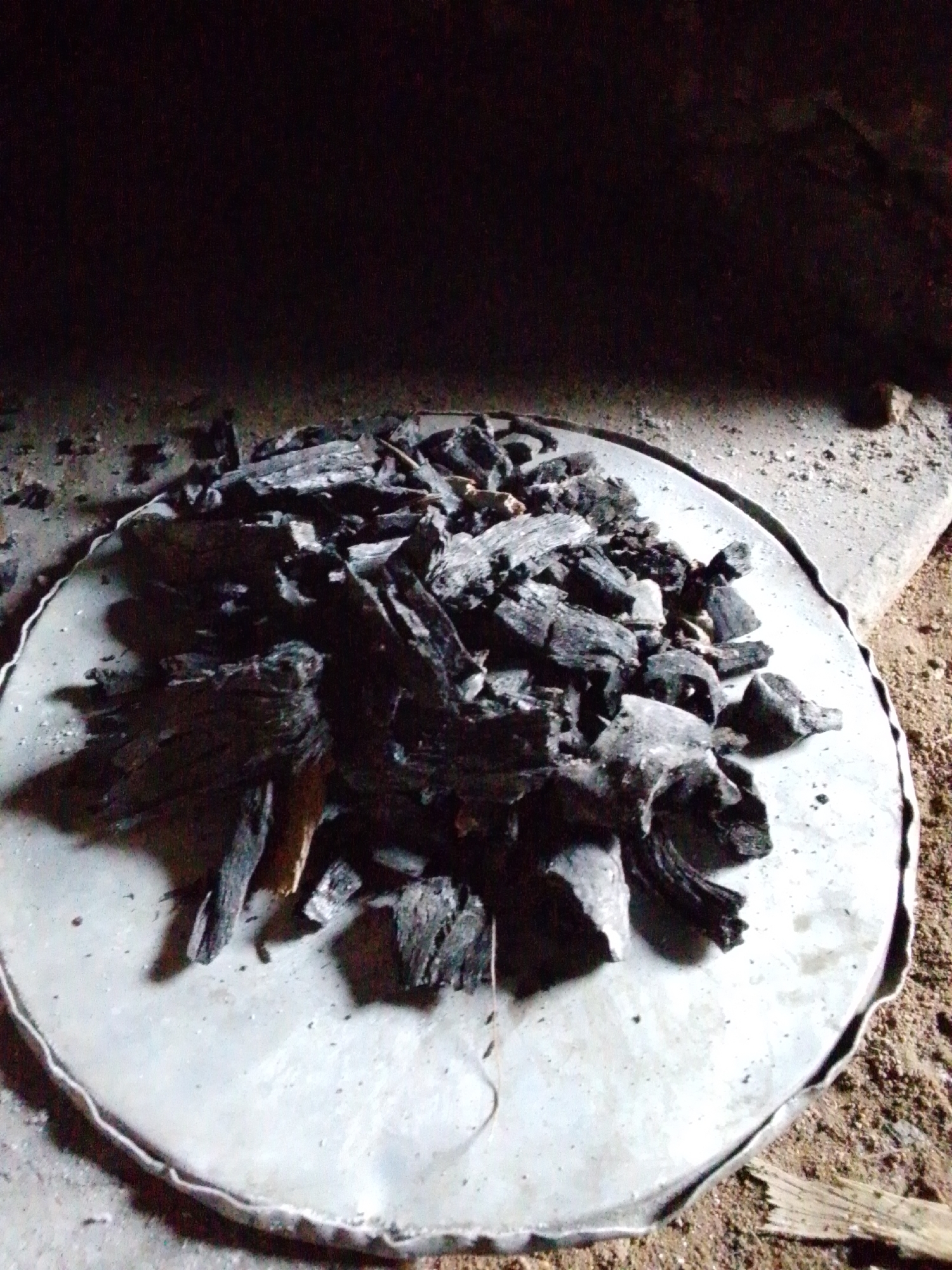 A close-up, medium-angle shot shows a pile of charcoal pieces on a round, gray metal tray. The tray has a raised, uneven edge and is placed on a surface composed of sand and what appears to be dried mud or clay. The background is dark and out of focus, suggesting an indoor or sheltered environment, possibly a kitchen or a storage area. There are no people visible in the image. The lighting is dim, casting shadows from the charcoal onto the tray. The scene is located in Jalingo, Nigeria.