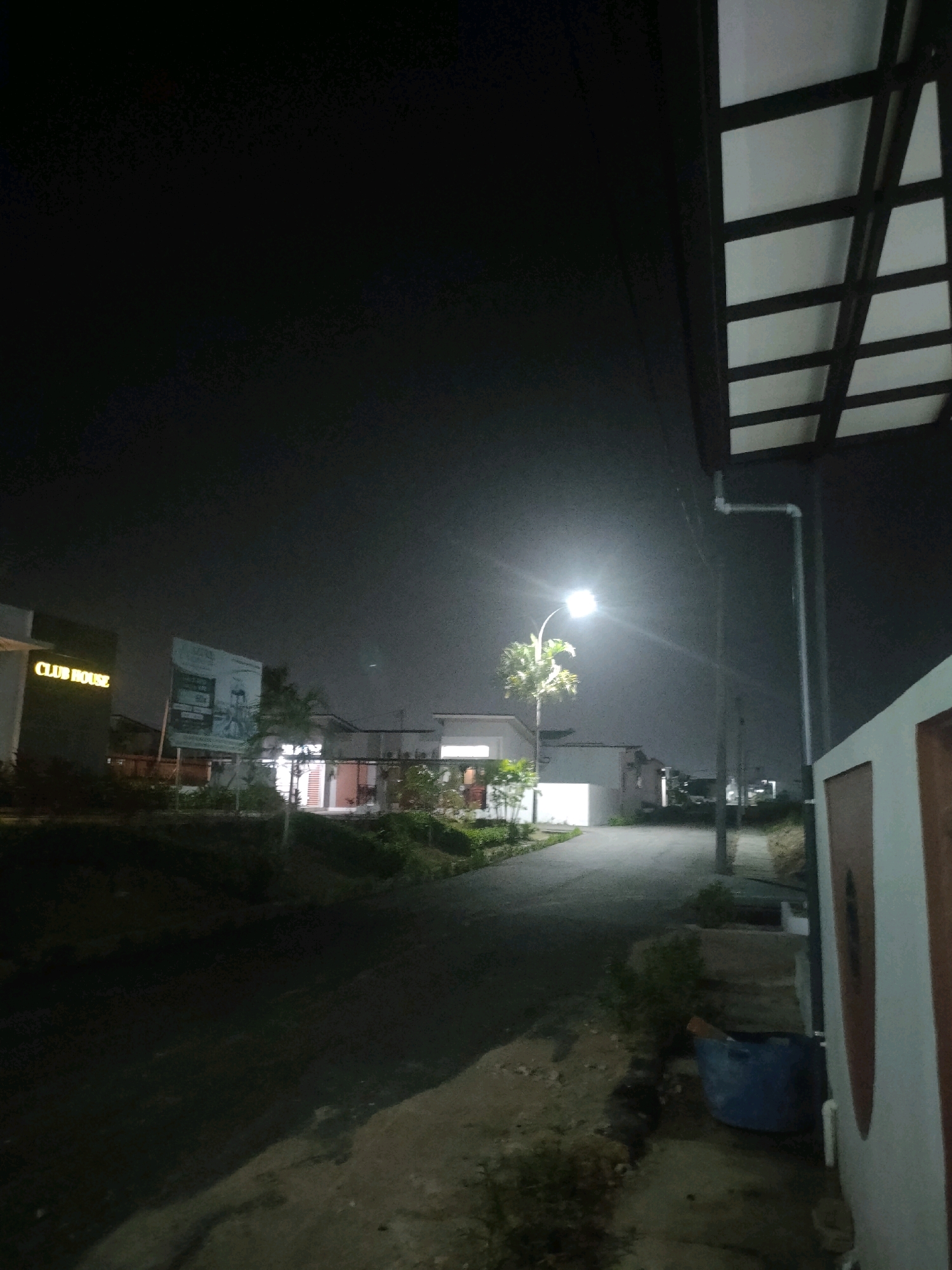 The image captures a street scene at night, illuminated by a street lamp. On the left, a building with a sign that reads "CLUB HOUSE" is visible. Adjacent to it, a large billboard is partially obscured by foliage. Further down the street, houses and other structures can be seen. In the foreground on the right, a portion of a building with a cream-colored wall and brown trim is visible. A blue trash can sits near the base of a pipe that runs down the exterior of this building. The overall impression is of a residential or community area in Batam, Indonesia, at night, with artificial lighting providing visibility.