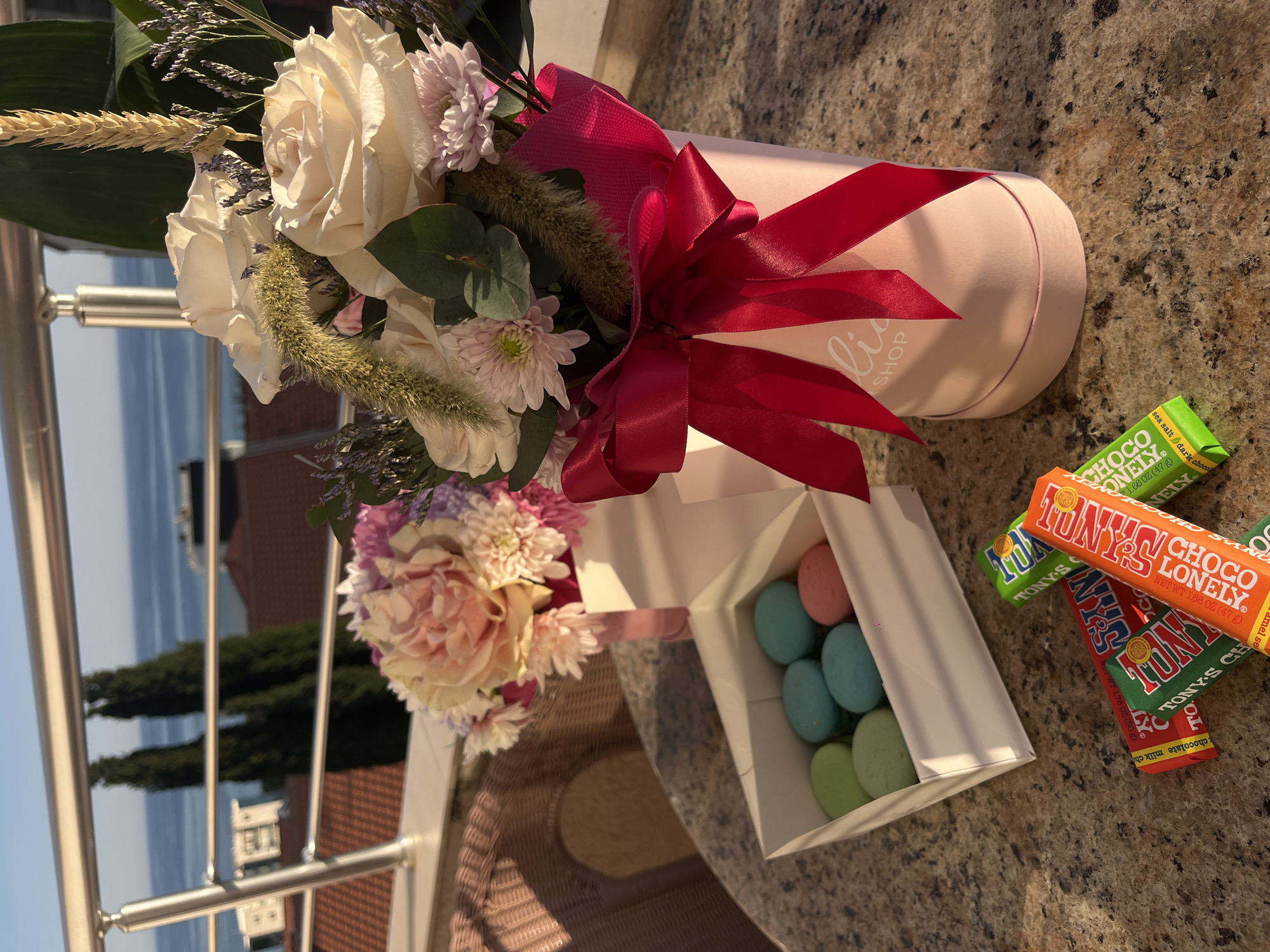 This media file presents a collection of celebratory items arranged on an outdoor surface, likely a balcony, overlooking a coastal scene in Bečići, Montenegro. The primary subjects include a bouquet of flowers, a box of macarons, and several chocolate bars.
The bouquet, nestled in a light pink cylindrical box, features a variety of blooms. Creamy white roses are prominent, accompanied by light pink and light purple chrysanthemums, delicate purple statice-like flowers, green foliage, and dried, feathery wheat-like elements, all tied with a vibrant dark pink ribbon. The cylindrical box has text "lia SHOP" visible on its side.
Adjacent to the bouquet is an open white box containing a selection of pastel-colored macarons in hues of light blue, pink, and green. Scattered nearby are several Tony's Chocolonely chocolate bars in various flavors, identifiable by their colorful wrappers. Visible text on these bars includes "TONY'S CHOCOLONELY", "sea salt / dark choco" on a green wrapper, "caramel se" (likely caramel sea salt) on an orange wrapper, and "chocolate milk ch" (chocolate milk chunk) on a red wrapper, all indicating "NET WT 1.66 OZ (47 g)".
The items rest on a mottled brown and cream surface, possibly a stone countertop or tiled floor. In the background, a metal balcony railing is visible, beyond which lies a panoramic view. The bright daylight suggests a sunny day. In the distance, the serene blue sea stretches out, flanked by buildings and lush green trees on the land, characteristic of a scenic coastal location like Bečići, Montenegro. The overall arrangement conveys a sense of thoughtfulness and celebration.