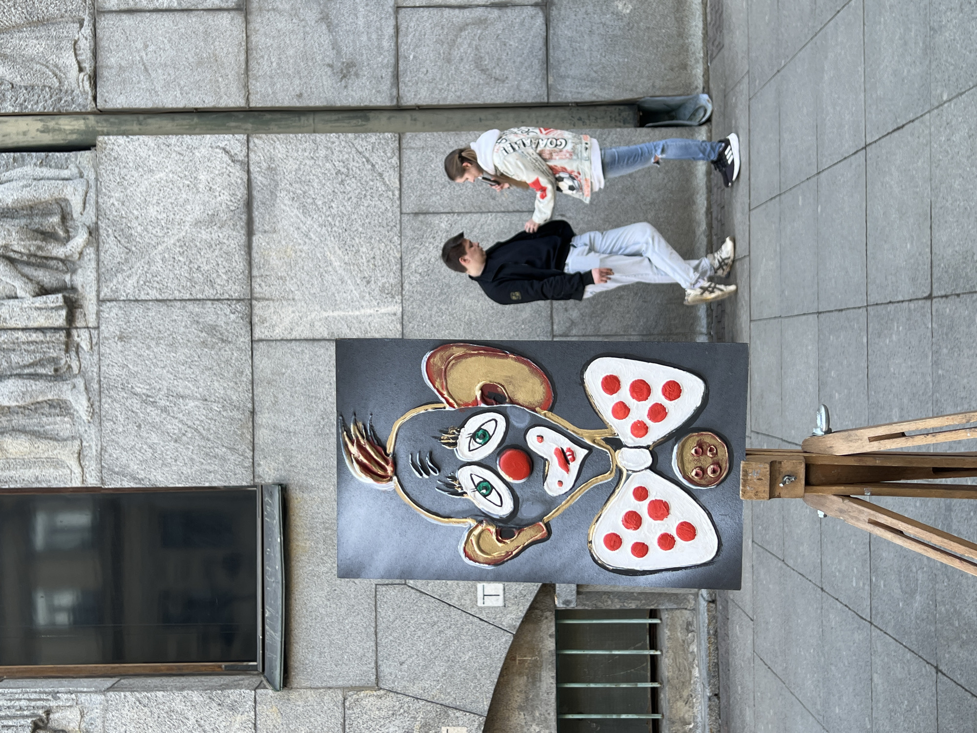 The image captures an urban street scene on a paved sidewalk in Saint Petersburg, Russia, against the facade of a large stone building.
In the immediate foreground, positioned slightly left of center, a vertical painting of a stylized clown's head and upper torso rests on a light brown wooden easel. The painting, primarily dark gray, features a textured surface depicting a clown with large golden ears, spiky golden-red hair, white eyebrows, green eyes with white pupils, a prominent red spherical nose, white lips forming a smile, and a white bow tie adorned with red polka dots. Below the bow tie is a gold circular button with four red studs.
To the right of the painting, two young adult individuals stand on the gray tiled sidewalk, facing each other. The male, on the left, wears a black zip-up jacket over a black t-shirt, light blue jeans, and white and beige sneakers. He is looking down. The female, on the right, wears a white hooded jacket with various colorful graphic prints, including "GOAAAAL!!", a soccer ball, and a distressed flag-like design in red, white, and blue. She has light blue ripped jeans and black athletic shoes with white soles. Her light brown hair is pulled back. She is holding a dark, rectangular object (possibly a smartphone) in her right hand, pointing it towards the male's midsection, specifically his jacket.
The background consists of a multi-story building with a facade constructed from large, light gray, rough-textured granite blocks. On the upper left, a dark rectangular window with a wooden frame and stone sill reflects interior elements. Above the individuals, a section of relief carving depicting the lower torsos and feet of multiple figures is visible on the stone facade. A thin, vertical, weathered green pipe extends down the right side of the building. On the lower left, a ground-level window or vent is covered by a green metal grate composed of vertical bars, with a sloped stone element above it.