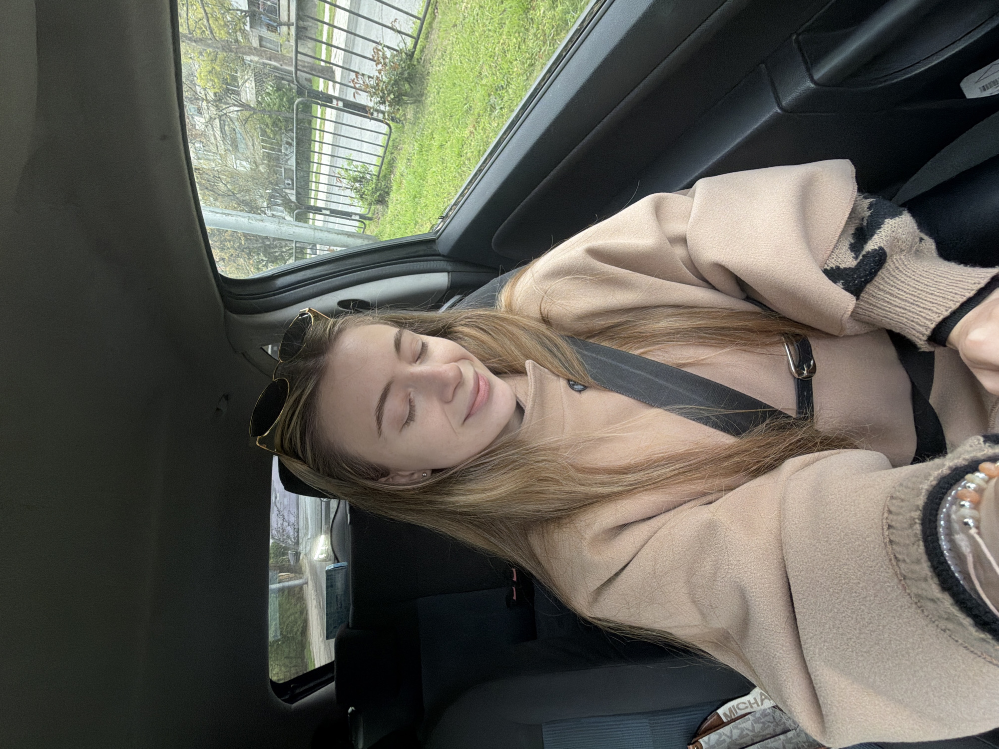 A young woman is seated in a car, wearing a beige coat and a black seatbelt. She has long, light brown hair and her eyes are closed. Sunglasses are perched on her head. Outside the car window, a street with a metal fence, trees, and grass can be seen. The car appears to be stationary. It is not possible to identify the exact location within Plovdiv based on the provided image.