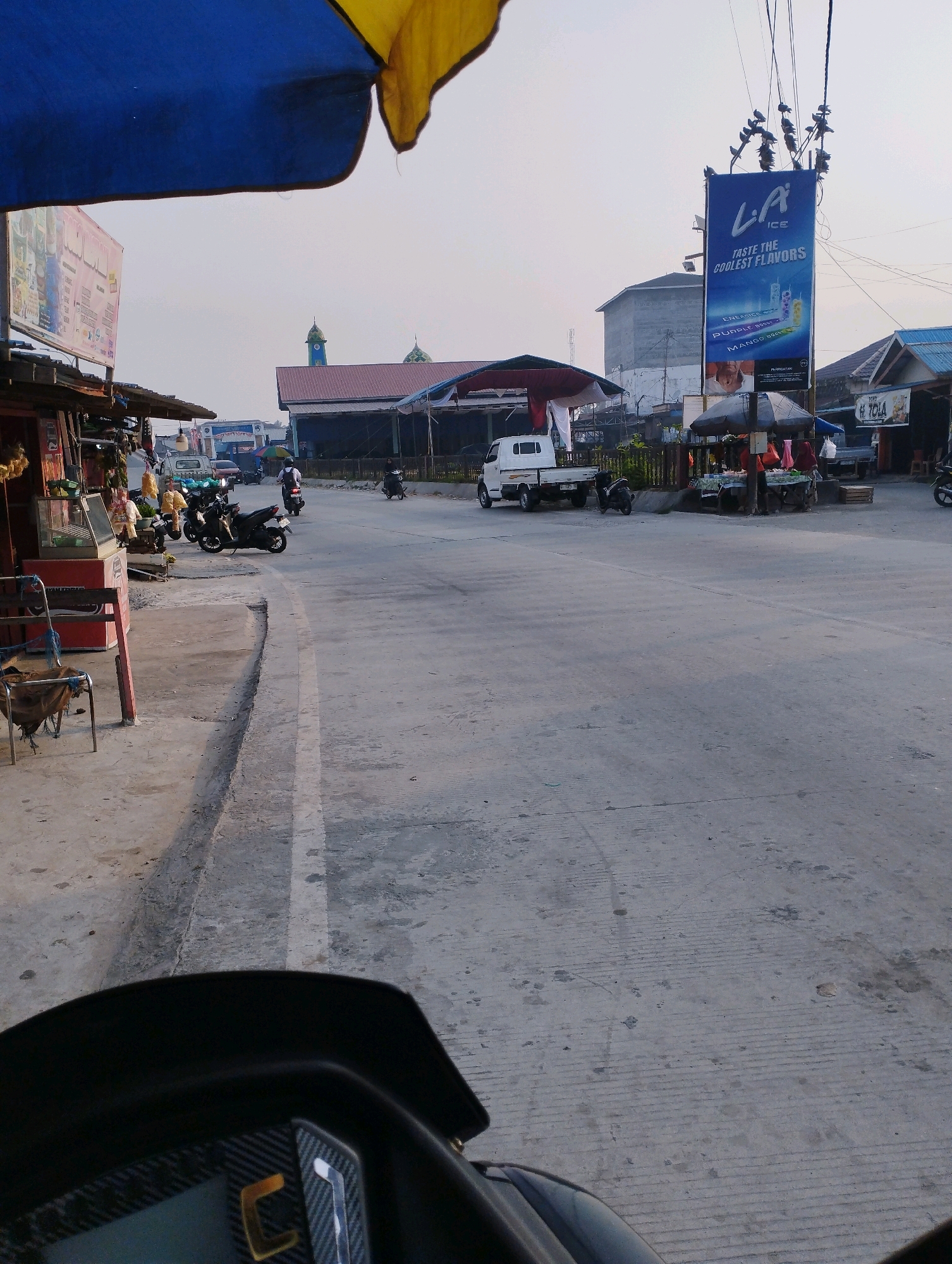 The image depicts a street scene in Kuala Samboja, Indonesia, under a bright, overcast sky. In the foreground, the handlebars and part of the dashboard of a black motorcycle are visible, indicating the photo was taken from the rider's perspective.
To the left of the road, a small roadside market is set up. Stalls with awnings display various goods, including what appear to be dried goods, food items, and possibly clothing. A glass display case is prominent in one of the stalls. A metal chair with a torn, burlap-like seat is positioned in front of one stall.
Across the road, a white flatbed truck is parked, and several motorcycles are also present. A large blue and yellow tarp extends over a section of the market area, providing shade. In the background, a billboard advertises "LA ICE" with the tagline "TASTE THE COOLEST FLAVORS." A tall, grey building stands behind the billboard, and further in the distance, the domed roofs of what appear to be religious buildings, possibly mosques, are visible.
The road itself is made of concrete and shows signs of wear, with painted white lines marking lanes. Several people are present, some standing at the market stalls, and others riding motorcycles. The overall impression is a typical day in a bustling roadside commercial area in Indonesia.
