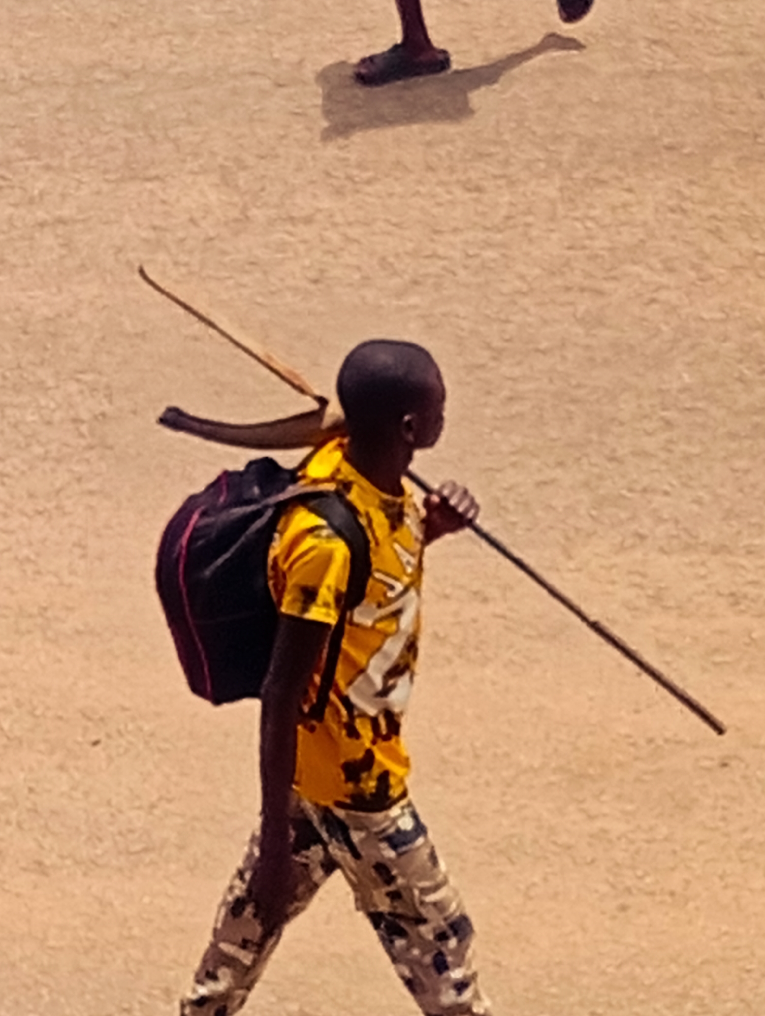 A young person is walking on a sandy-colored surface. They are wearing a yellow patterned shirt, camouflage-patterned pants, and a black backpack. A long, thin stick is held in their right hand, resting on their shoulder. A bow and arrow are positioned behind the backpack, also resting on the shoulder. In the upper part of the frame, there are feet walking.
The location is the city of Bukuru, Nigeria.