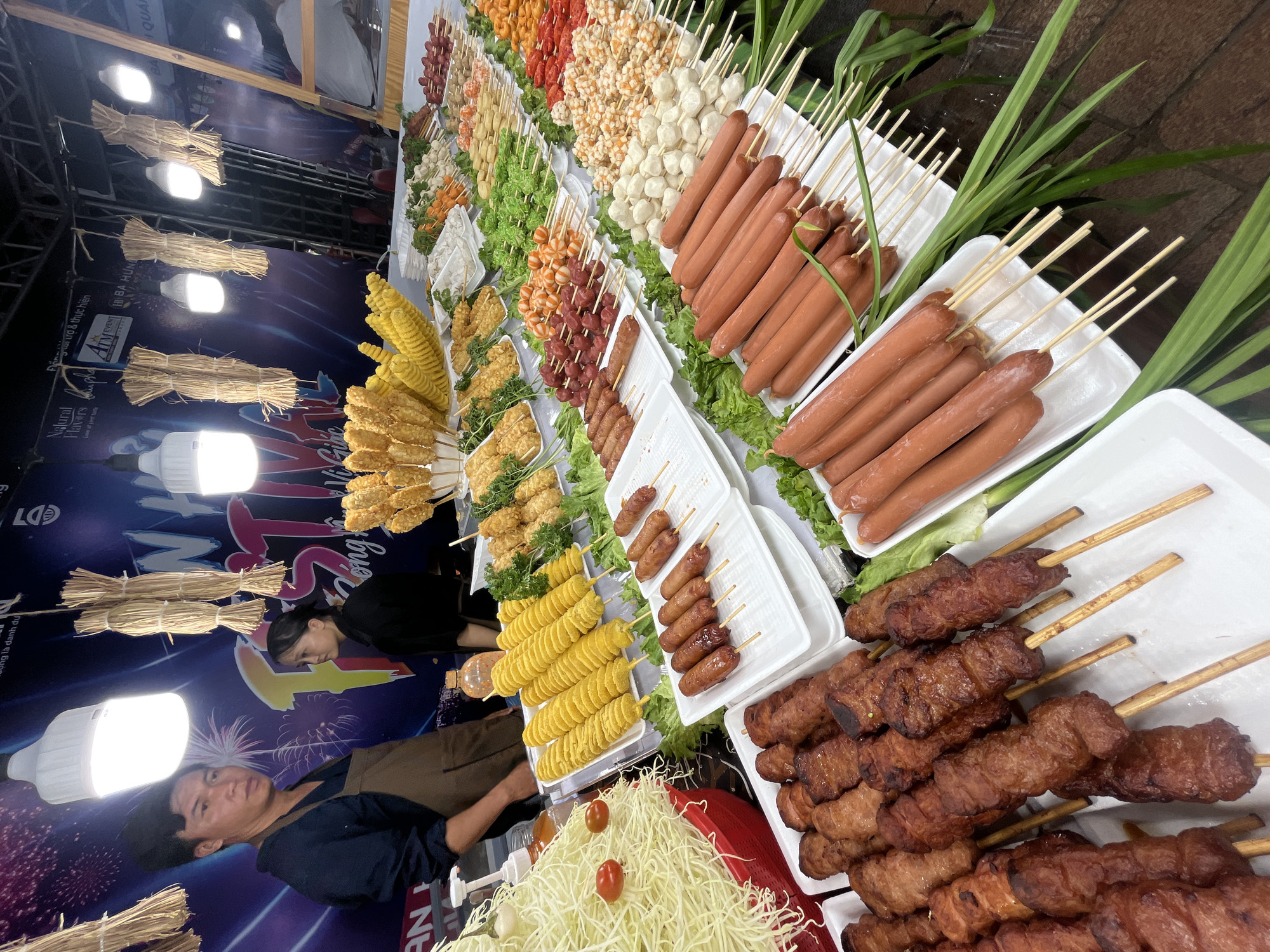 This image depicts a vibrant street food stall in Da Nang, Vietnam, likely operating during a festival. A male vendor, wearing a brown apron over a dark shirt, stands to the left, his gaze directed forward. Beside him, a female vendor in a black shirt looks to her right. The stall is laden with a diverse array of meticulously displayed food items, primarily skewered and fried.
Dominating the foreground are skewers of long, reddish-brown sausages and what appear to be grilled meat pieces. Behind these, rows of smaller, round meatballs and other processed meat items are arranged on skewers. Further back, there are skewers of battered and fried items, some resembling corn on the cob, and spiral-cut potatoes. A large pile of shredded cabbage, garnished with two small tomatoes, sits in a red bowl on the far left.
The food is presented on white rectangular trays and is artfully arranged with fresh green lettuce as a base. Hanging above the stall are bright LED lights and bundles of straw, contributing to a festive atmosphere. The backdrop features a large banner with colorful graphics and text, including the word "FESTIVAL" prominently displayed, suggesting a special event. The overall scene captures the lively ambiance of street food culture in Da Nang, with an emphasis on variety and visual appeal.