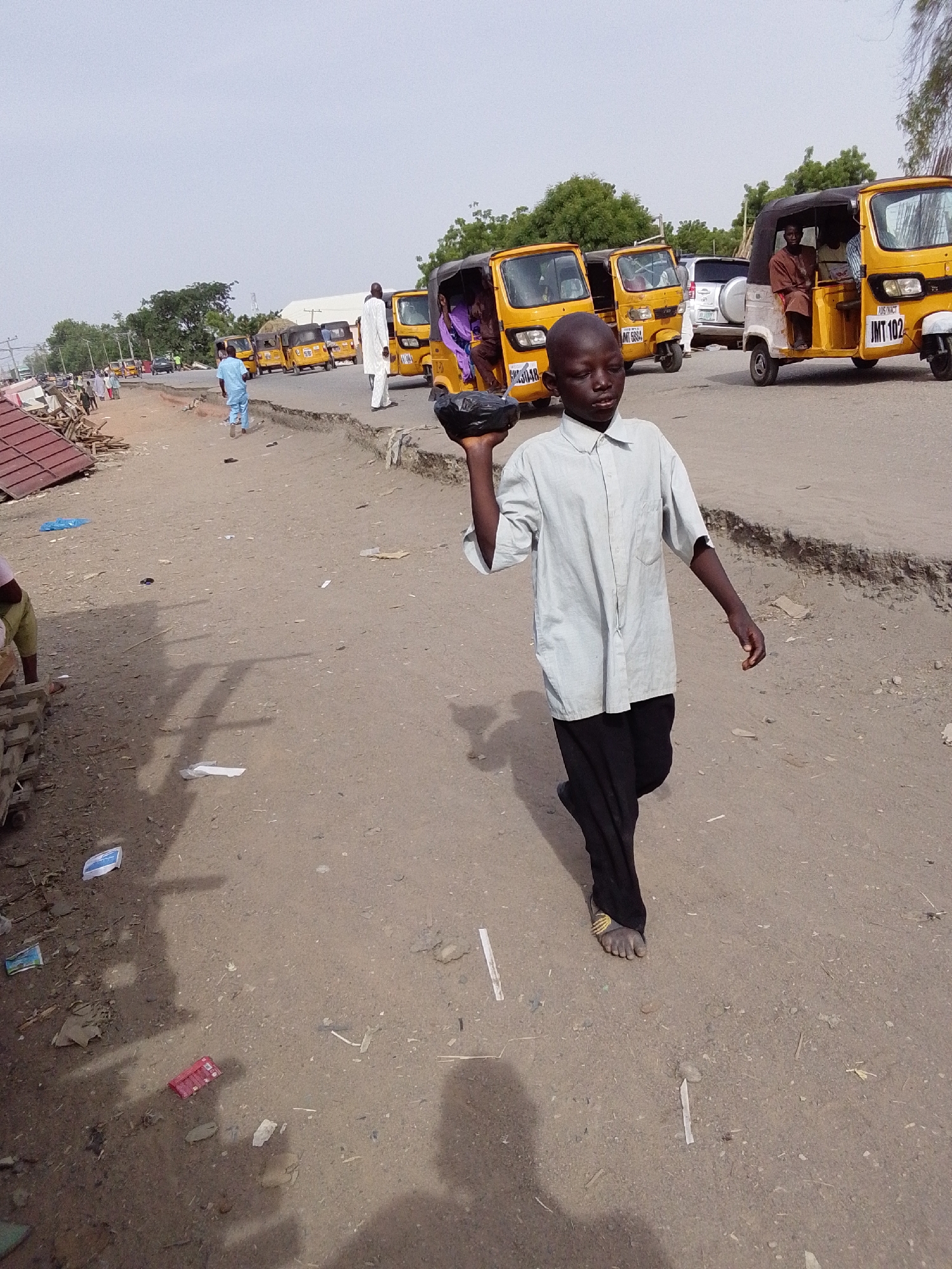 A young boy walks on a dirt road in Jimeta, Nigeria, carrying a black object in his right hand. He wears a light grey, short-sleeved shirt and dark pants, with sandals on his feet. Behind him, a line of yellow auto rickshaws (tuk-tuks) is parked along the side of the road. Other people are visible in the background, and debris litters the ground. The road surface is cracked and uneven.