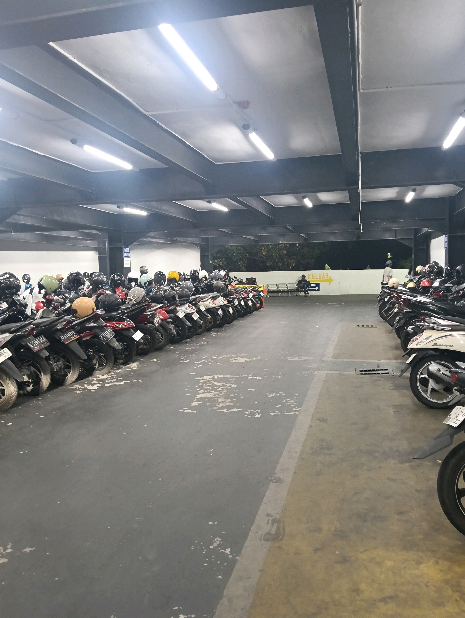 The image shows a packed motorcycle parking lot, likely at the Mal Olympic Garden (MOG) mall in Malang, Indonesia, given the presence of the "KELUAR" (Exit) sign. A long row of parked motorcycles extends from the foreground to the background on the left side of the image. The motorcycles are a variety of colors and models, with many displaying helmets on their seats or handlebars. The parking lot is covered by a dark ceiling with prominent fluorescent lights, indicating an indoor or semi-indoor space. The floor is concrete with some scuff marks and areas of discoloration. On the right side of the image, another section of the parking lot is visible with more motorcycles. In the background, a white wall with a yellow arrow sign pointing to the left and the word "KELUAR" is present, along with a person sitting on a bench and another person standing nearby. Trees and vegetation are visible beyond the wall. The overall impression is one of a busy and functional parking facility.