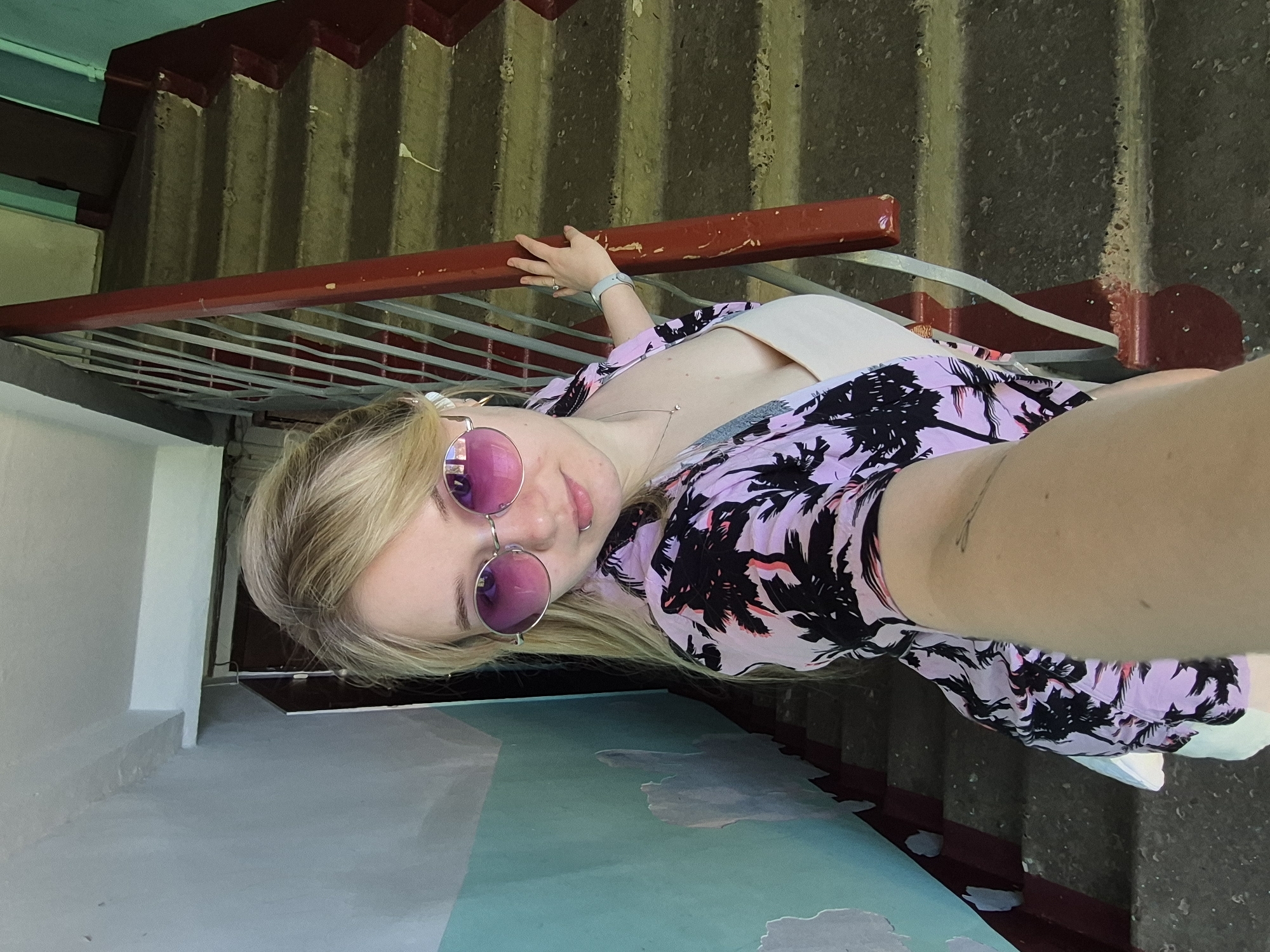 A blonde woman wearing pink sunglasses and a lavender and black palm tree print shirt stands on a staircase in what appears to be an entryway. She is holding onto a red railing with a metal grate beneath it. The staircase is painted in a color pattern with shades of green and blue in the foreground, and grey, textured concrete steps in the background. The building appears to be in Chaikovsky, Russia.
