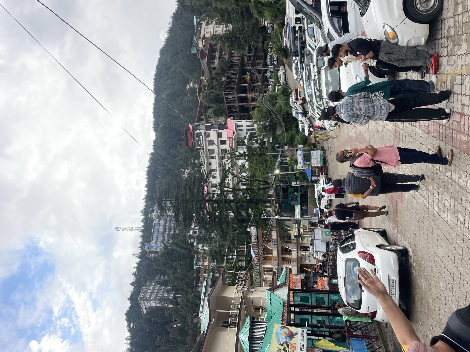 The image captures a street scene in Bhagsu, India, with a focus on a busy road lined with buildings and vehicles. In the foreground, a white Hyundai i20 hatchback, license plate PB10C06050, is parked on the right side of the road. Several people are present, including a group of young men standing near a white car, one of whom is on his phone. Another person, wearing a pink shirt and a backpack, is also in conversation on their phone. Further down the road, a man in a dark shirt and brown pants walks away from the camera.
The environment is a hilly urban landscape. Buildings, some multi-story with balconies and red roofs, are constructed on the steep slopes of a forested mountain that dominates the background. Pine trees are interspersed among the buildings and cover the hillside. Overhead, power lines stretch across the cloudy sky, and a tall communication tower is visible on the crest of the mountain. The road surface appears to be paved with interlocking bricks, and there are signs and shops along the street, suggesting a commercial area. The overall impression is one of a vibrant, bustling mountain town.