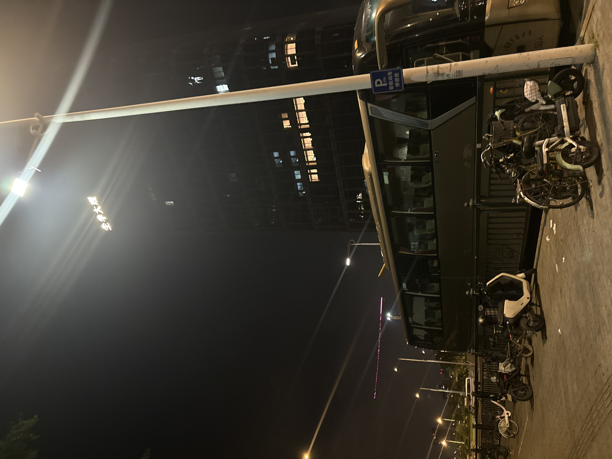 The image depicts a street scene at night in Haizhu District, China. The main subjects are a double-decker bus and several parked bicycles and electric scooters. The bus is dark-colored and appears to be at a bus stop, with its windows reflecting the surrounding lights. A blue and white sign with a "P" symbol is visible on the bus, indicating parking.
Numerous bicycles and electric scooters are neatly parked along the sidewalk next to the bus. These range from traditional bicycles to more modern electric models, some with baskets. The lighting is typical of a nighttime urban environment, with streetlights and illuminated windows from a nearby building casting a glow.
The building in the background is a tall structure with many windows, some of which are lit, suggesting it is an office building or residential complex. The overall scene is one of a quiet, urban night, possibly a transportation hub or a street with a bus stop. There are no people visible in the image. The weather appears clear.
The text visible on the blue sign translates to "Parking," with additional Chinese characters below that likely specify parking regulations or information. Another sign on the bus is partially visible and appears to have Chinese characters as well, possibly indicating the bus route or destination.