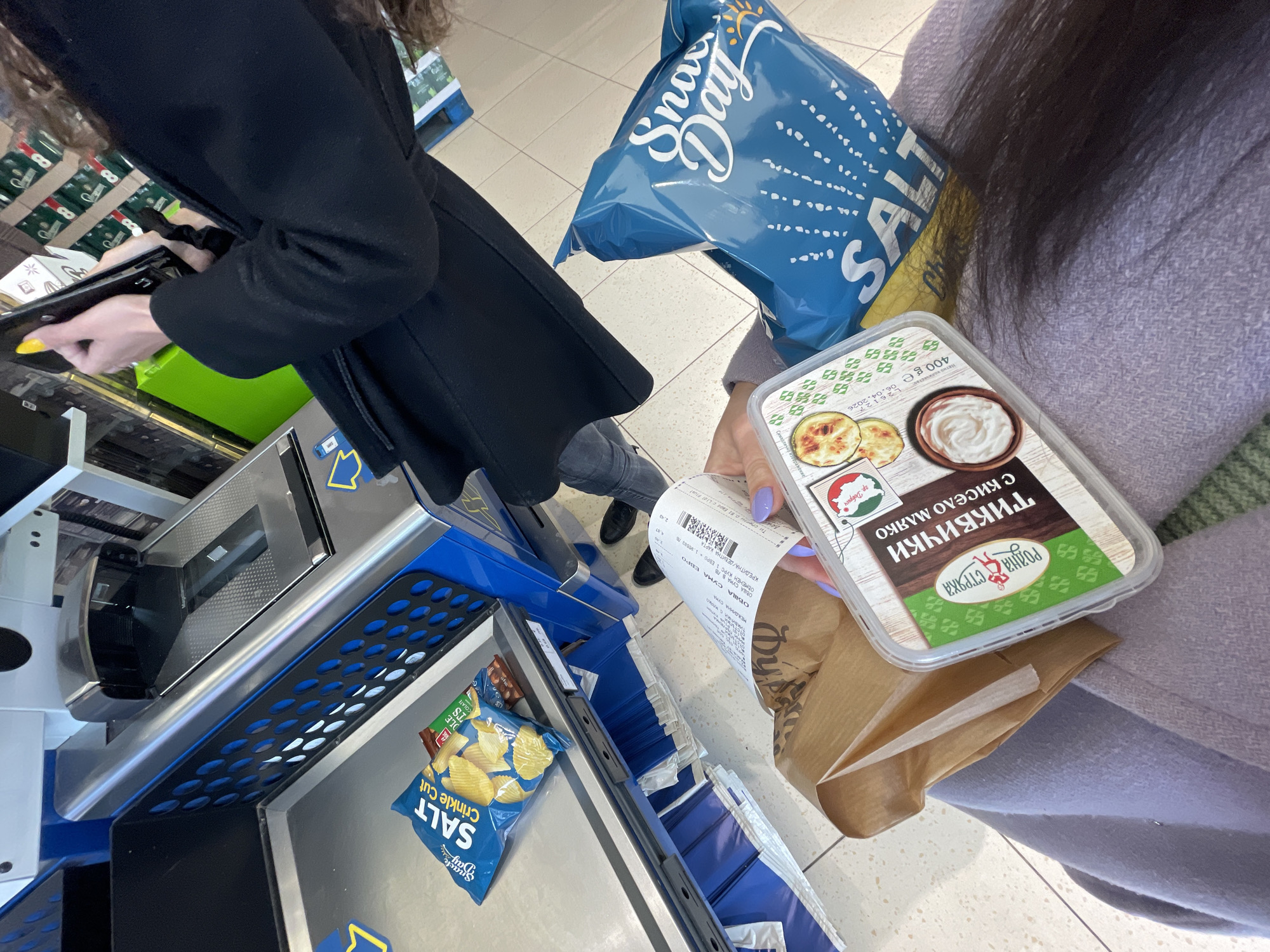 The image shows a person at a self-checkout in a supermarket in Sofia, Bulgaria. The self-checkout is blue and silver. The person is scanning items, including a bag of "Snack Day" salt and vinegar chips and a container of "Тиквички с кисело мляко" (zucchini with yogurt) from "Родна стряка" (Rodna Stryaka) made in Dobrich. The person is also holding a brown paper bag and a receipt. The person has purple nail polish. Another person in a black coat is visible to the left, scanning their items. The supermarket has stacks of merchandise visible in the background.