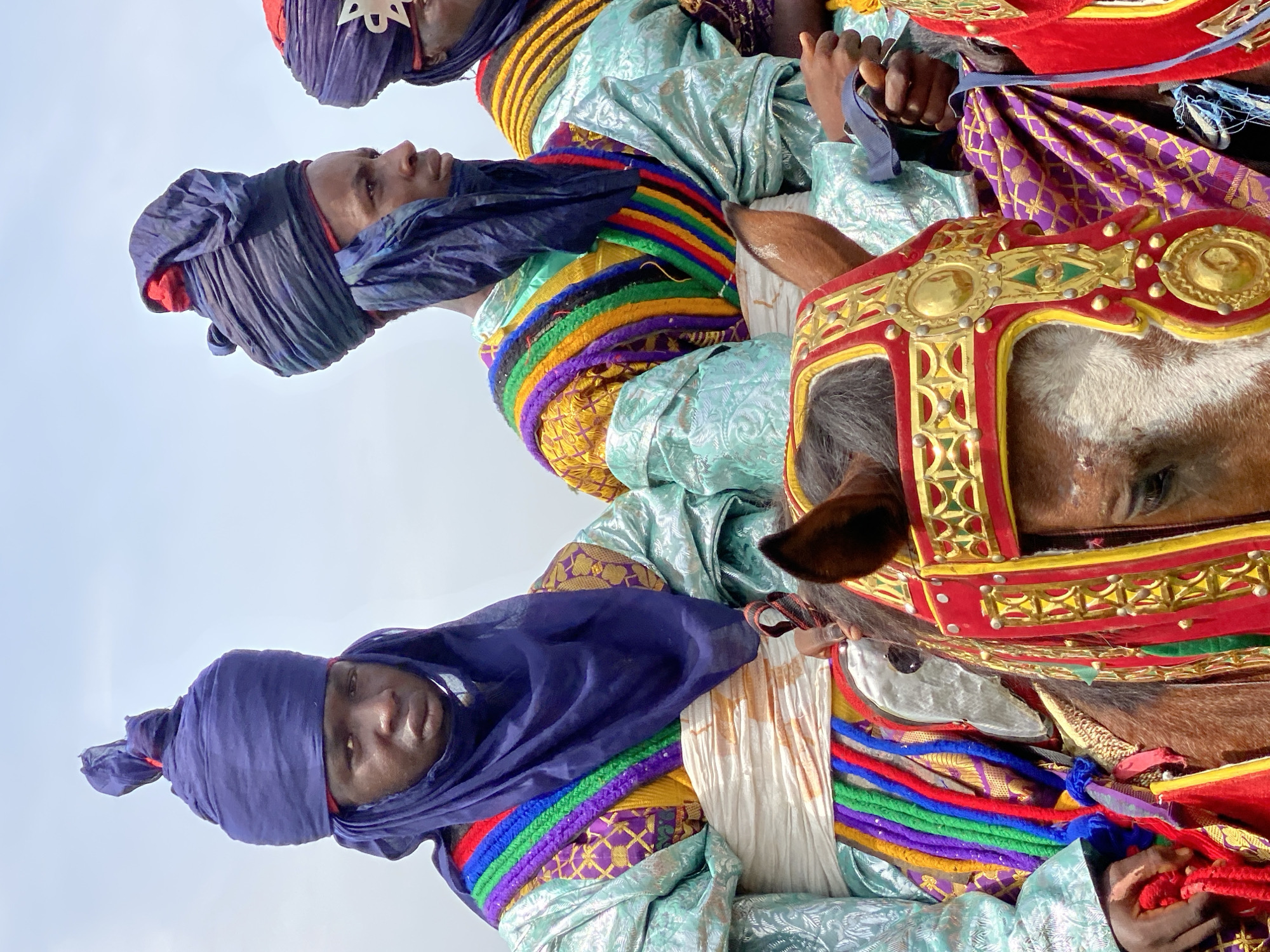 The image depicts three men on horseback in Kafanchan A, Nigeria. Each man wears a dark blue turban, a light blue, patterned robe, and a colorful rope around their chest. The horses are elaborately decorated with red and gold headgear. The background is a pale sky.
