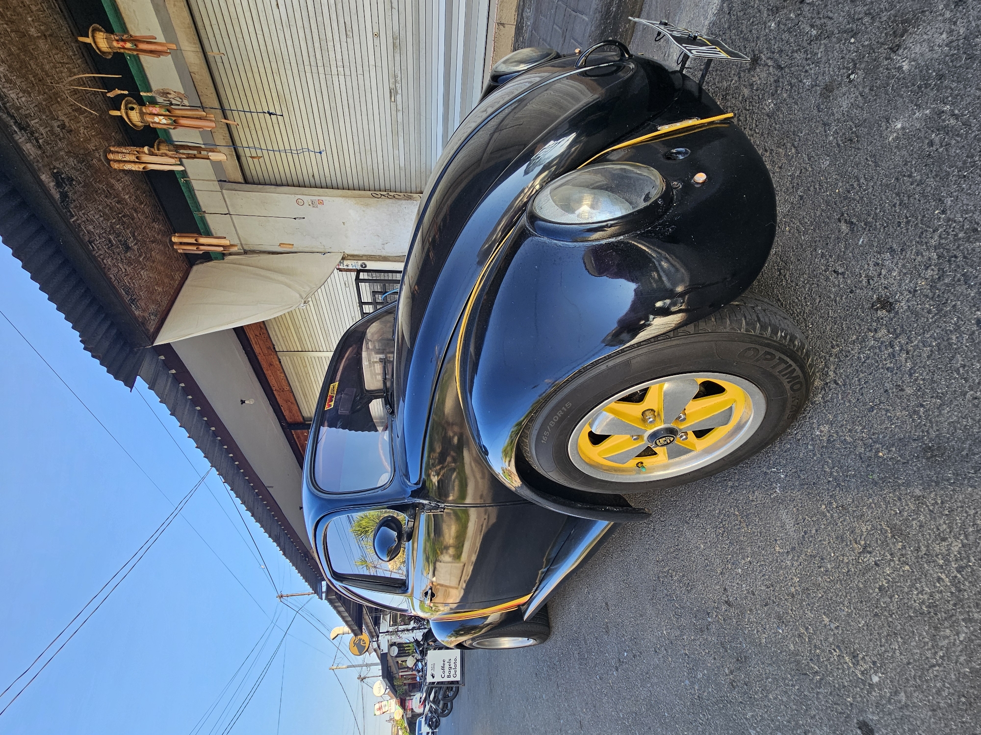 An eye-level, low-angle, full shot captures a black Volkswagen Beetle parked on an asphalt street in Denpasar, Indonesia. The car, positioned at an angle, faces slightly towards the right. Its front fender is adorned with a yellow stripe that extends along the side of the vehicle. The car features bright yellow rims on its wheels, with a black tire displaying the text "165/80R15". A yellow stripe also runs along the side of the car. In the background, a building with a corrugated metal roof is visible, with roller doors that appear to be closed. Bamboo wind chimes hang from the eaves of the building. A white awning extends from the building, and a sign with the text "Coffee Bagels. Gelato." is visible on the left side of the image. Several motorbikes are parked in front of the building. The sky above is clear and blue.