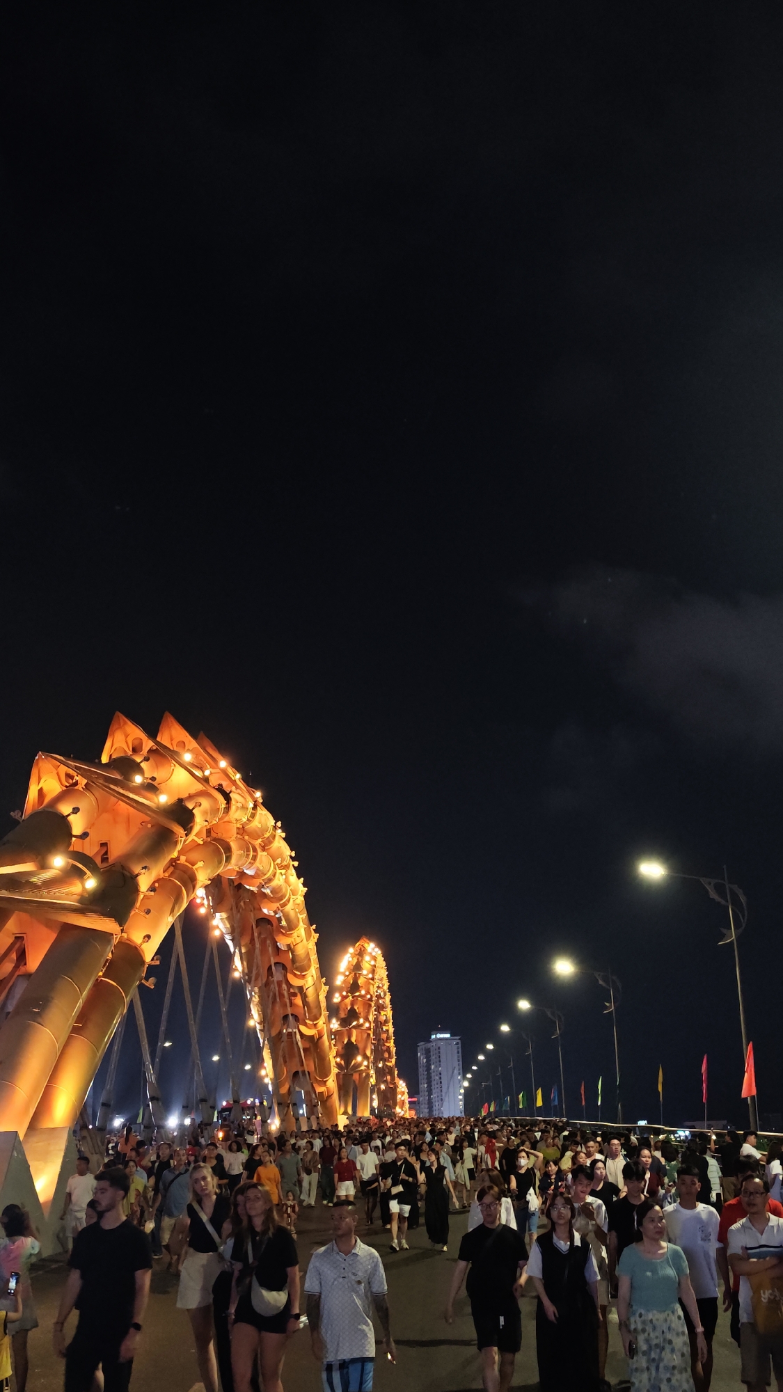 Captured at night in Đà Nẵng, Vietnam, the scene unfolds on a bridge illuminated by bright orange lights that form the dragon-shaped arch of the Cầu Rồng (Dragon Bridge). The bridge is crowded with people walking, creating a dynamic flow amidst the urban setting. Streetlights cast a glow along the bridge's edge, interspersed with colorful flags fluttering in the background. A tall building is visible in the distance beyond the bridge. The night sky forms a dark backdrop, partially obscured by clouds.