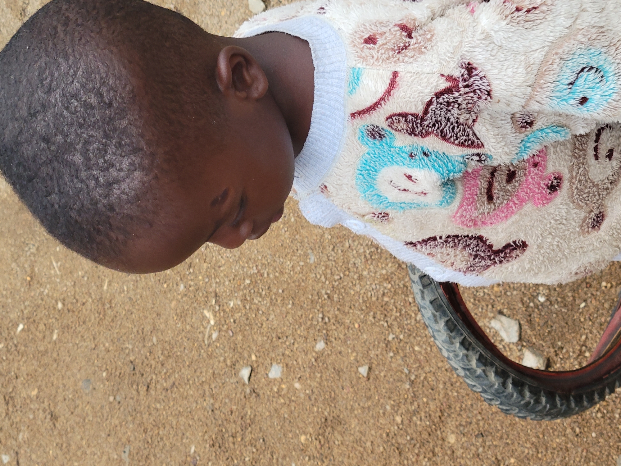 The image presents a top-down view of a young child, likely a baby or toddler, lying on a dusty, gravelly ground surface. The child has dark skin and short, dark hair, with eyes appearing closed or partially closed, suggesting they are resting or asleep. A small mark is visible on the child's forehead.
The child is wearing a soft, fuzzy garment, possibly a blanket or a pajama top, which is cream or off-white with a light grey/white ribbed collar. This garment is adorned with colorful patterns, including blue, pink, and reddish-brown figures that resemble cartoon animals or teddy bears.
In the bottom right corner of the frame, a segment of a bicycle wheel is visible, featuring a dark, textured tire and a reddish rim, resting on the ground. The ground itself is light brown, consisting of fine dirt and small stones, characteristic of an unpaved outdoor area.
The scene is brightly lit, indicating daytime. The child appears peaceful. There is no visible text in the image. The context places this scene in Jos, Nigeria.