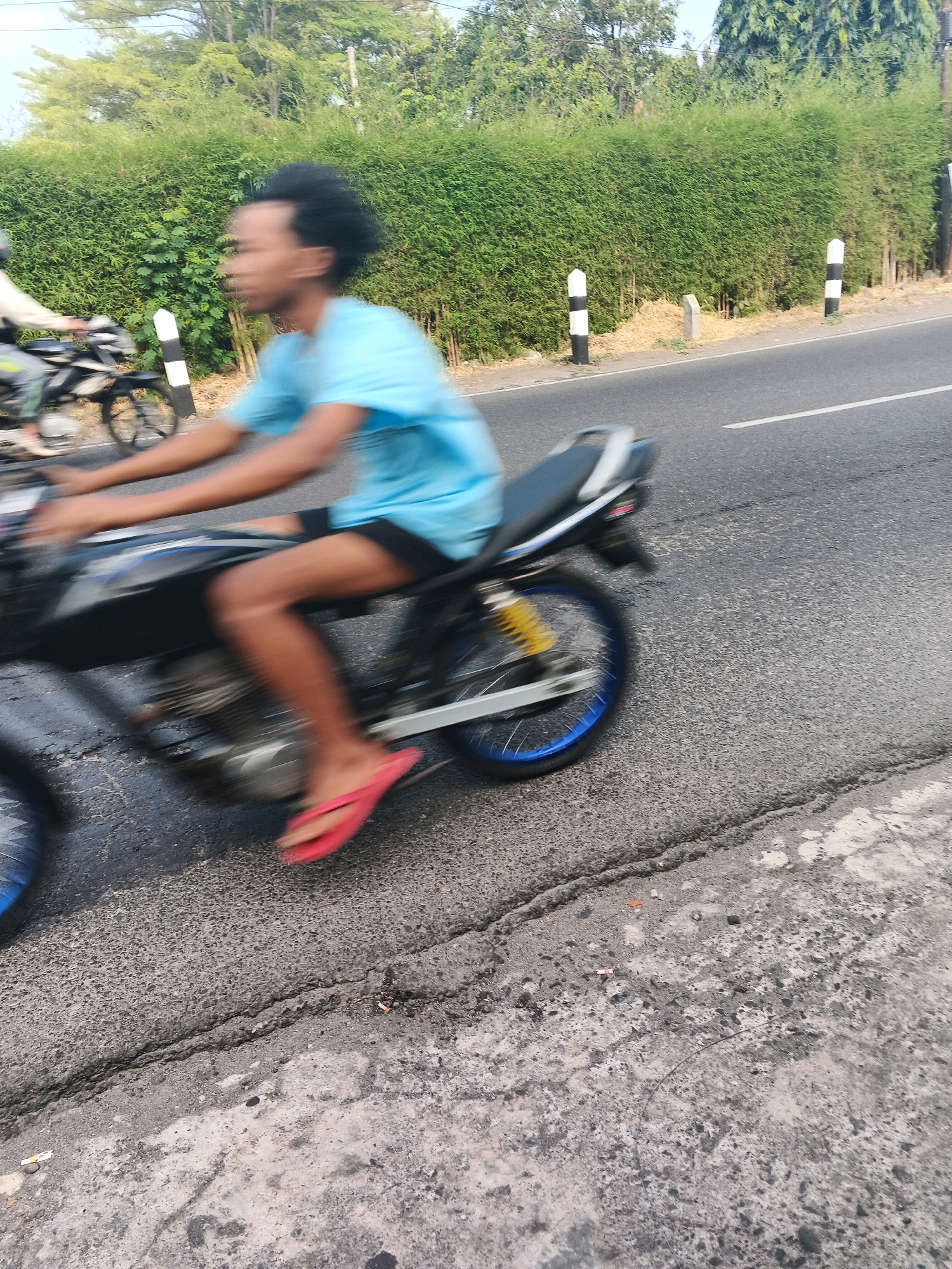 A person is riding a motorcycle on a road in Ungaran, Indonesia. The person is wearing a blue t-shirt and red flip-flops. The motorcycle is black and has blue rims. A dense green hedge runs along the side of the road, with two black and white striped posts visible in front of it. The road is paved with asphalt and appears to be cracked in places. The sky is visible in the background, and there are trees and other vegetation. The image is blurry due to motion, indicating the motorcycle is in motion.