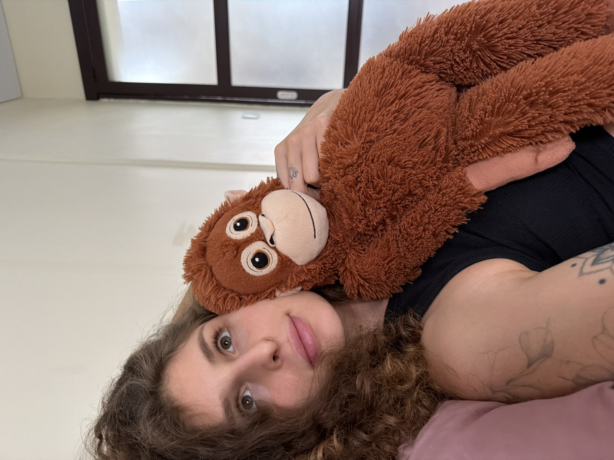 A young woman with curly brown hair is lying down, holding a brown plush orangutan. She is looking up and to the left with her eyes wide open. She is wearing a black tank top. The orangutan is positioned next to her face, with its arm wrapped around her shoulder. Her left arm is visible, showing tattoos on her forearm and shoulder. She is on a pink pillow. The background consists of a plain wall and a door with glass panels. The scene appears to be indoors, likely in a bedroom or living space. There is no indication of the specific location in North Kuta, Indonesia.