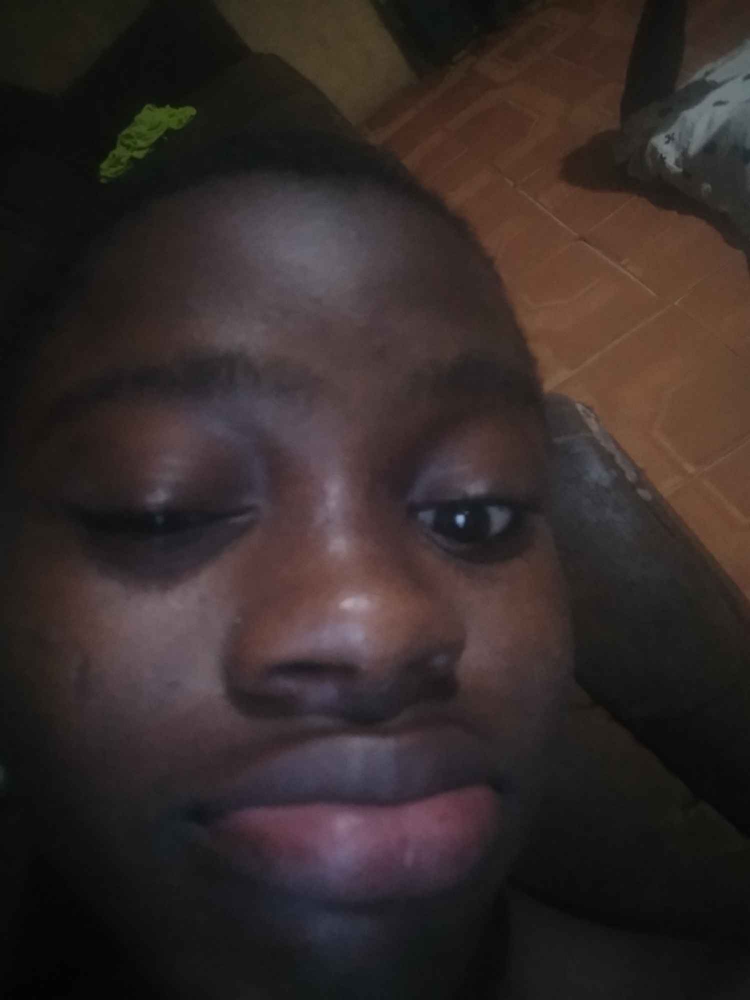 A close-up, slightly angled selfie of a dark-skinned person with dark hair is shown. The person is partially winking, with one eye open and the other mostly closed. Their lips are slightly pursed. The background is out of focus, showing a dark-colored object (likely a piece of furniture) and a light-colored floor. A small green object is located at the top of the image. The location is Unknown, Unknown.