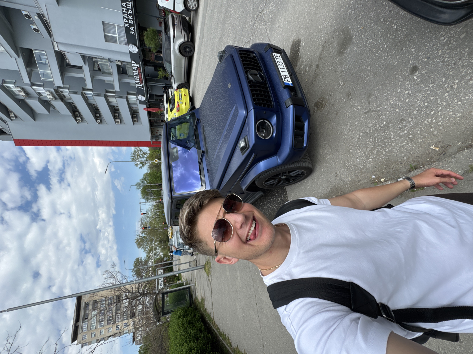 The image, rotated 90 degrees clockwise, captures a cheerful man taking a selfie on a city street. The man, fair-skinned with short brown hair and sunglasses, wears a white t-shirt, a gold chain, and a watch, with a black backpack strap visible. He smiles widely, showing his teeth.
Behind the man, a distinctive matte blue Mercedes-Benz G-Wagen is prominently parked. Its Bulgarian license plate "PB 1133 TE" is clearly visible, indicating the location is Plovdiv. To the left of the G-Wagen, a yellow car and a silver SUV are partially visible, alongside another white car further back.
In the background, a multi-story grey building with numerous air conditioning units on its facade stands to the left. A sign on this building reads "ДАНМАР" (DANMAR) and "ХРАНА ЗА ВКЪЩИ!" (FOOD FOR HOME! / TAKEAWAY FOOD!). Further down the street on the right, another multi-story building with many windows is visible. The scene includes asphalt pavement, a sidewalk, streetlights, and some greenery.
The time of day appears to be daytime, with bright light and a cloudy sky. The man's broad smile suggests a happy or content mood. The overall setting is an urban environment in Plovdiv, Bulgaria.