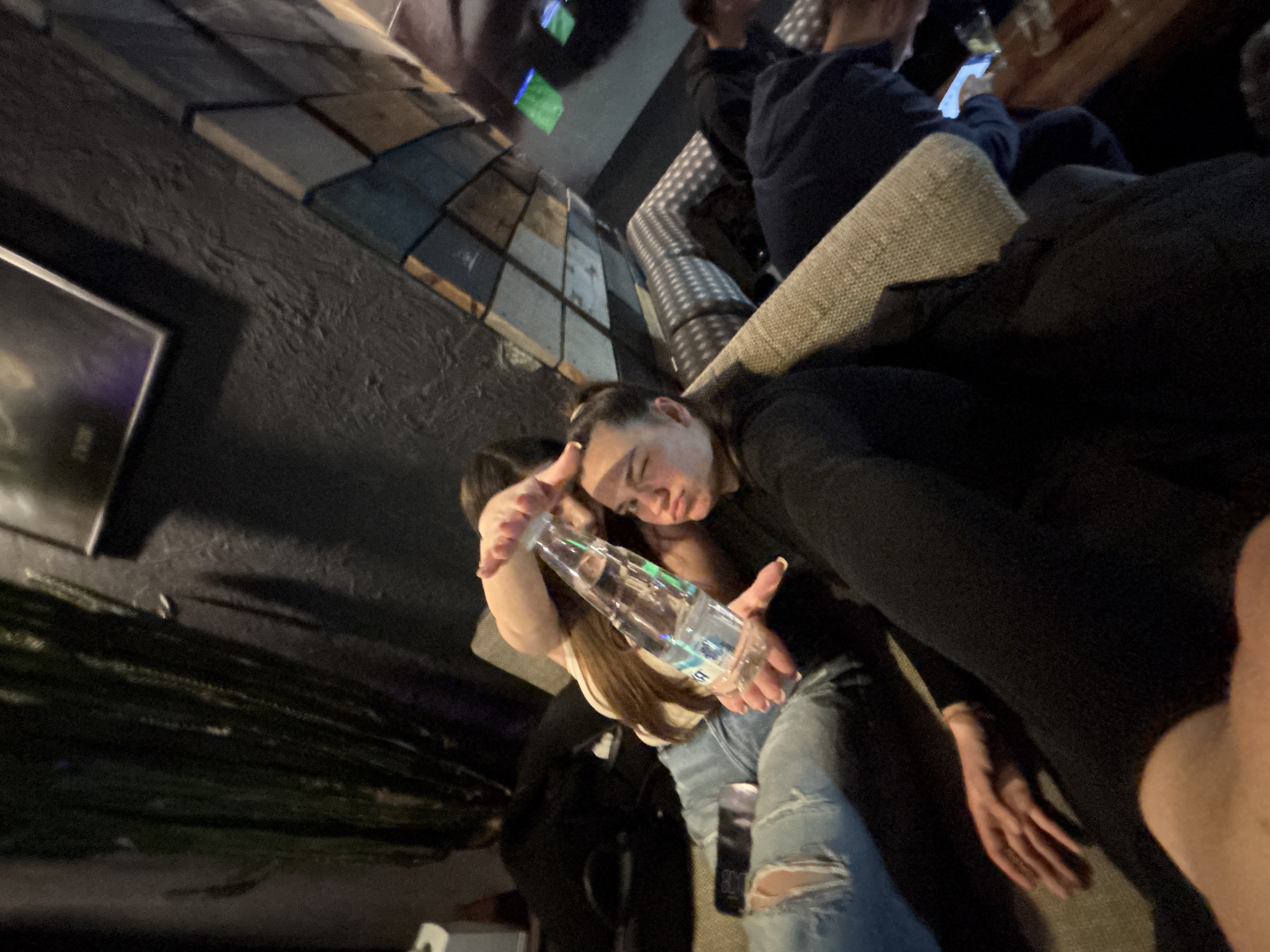 The scene is indoors, likely a bar or lounge in Plovdiv, Bulgaria. Two young women are the primary subjects, one holding a plastic water bottle and gesturing with her hand. The woman is wearing ripped jeans and a light-colored top. The other woman is partially obscured, and they appear to be embracing or posing together. Other people are visible in the background, sitting on a couch, with one appearing to use a mobile phone. The background includes a wall with patterned wooden tiles and a framed picture. The lighting is dim. A decorative green plant is present. The composition is from a high angle, slightly tilted.
