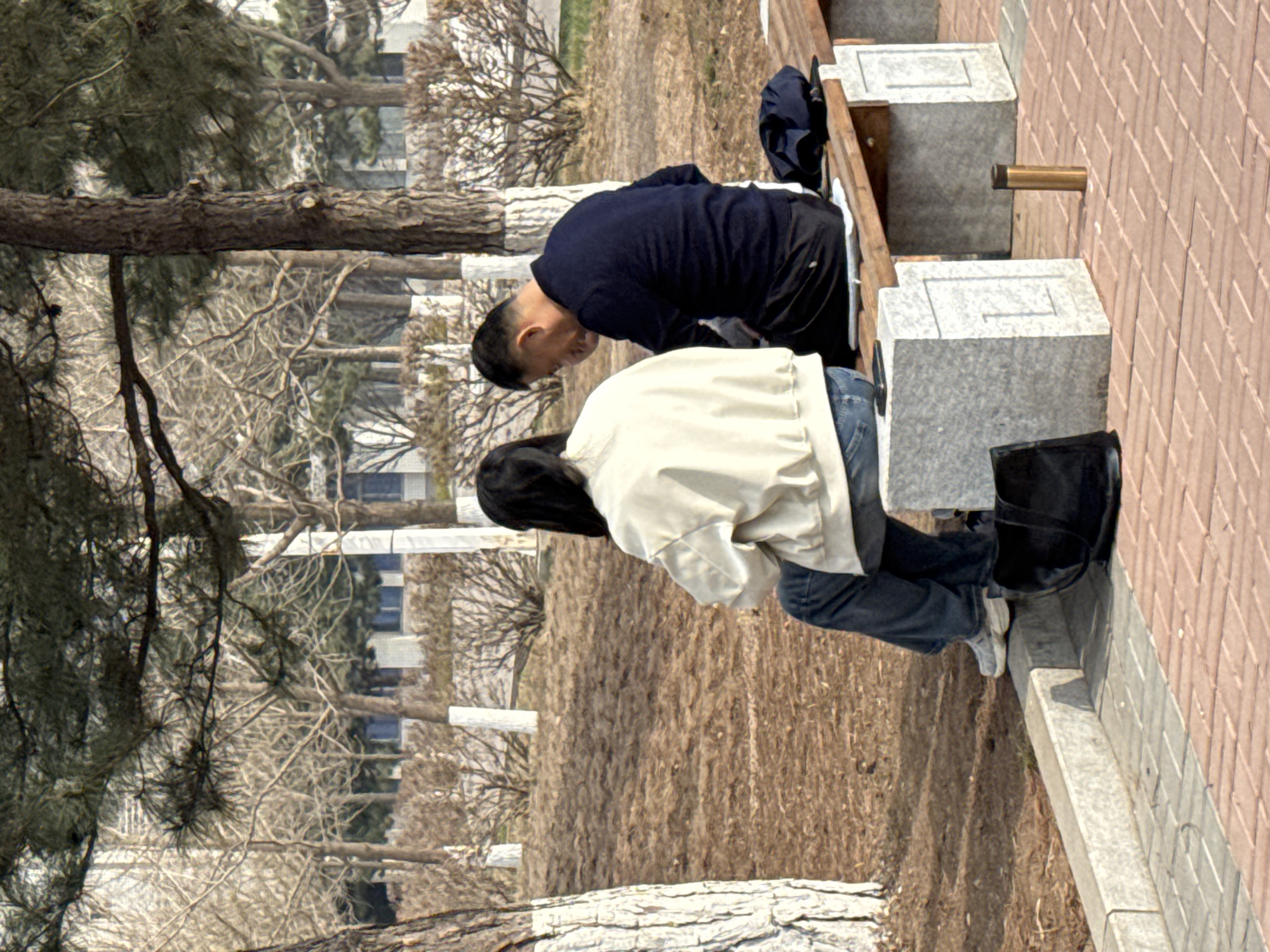 Two individuals, an older man and a younger woman, are seated on a bench in an outdoor location in Zhongtun, China. The man is wearing a dark blue long-sleeved shirt and dark pants. The woman is wearing a light beige jacket and blue jeans, with dark boots placed beside her. The bench is constructed of gray stone blocks. The background consists of bare trees, a grassy area, and distant buildings, suggesting an urban park setting in Zhongtun, China. A pipe is mounted on the brick wall behind the bench.