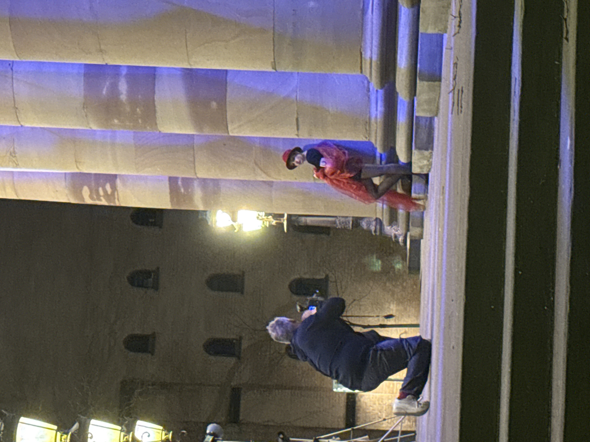A photographer takes a picture of a person on the steps of the Palais Rohan in Strasbourg, France. The photographer kneels on a platform and points the camera at the subject, who is wearing a red hat and a red shawl. The building facade is visible behind the photographer and the columns of the Palais Rohan are visible behind the subject. The scene is lit by street lights and the Palais Rohan.
