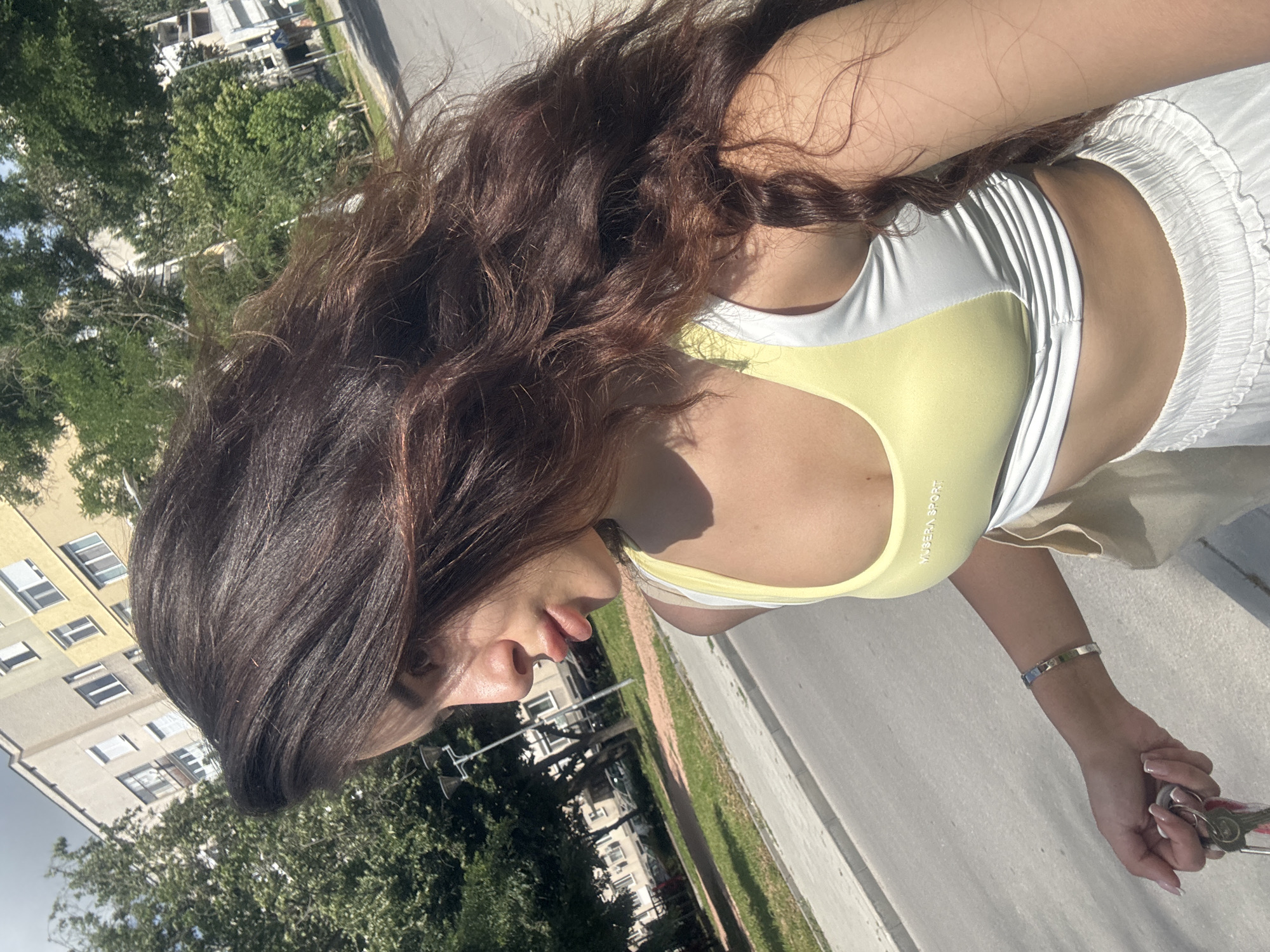 A young woman with long, dark, wavy hair is photographed outdoors, in a city setting. She is facing to the left, with her head turned slightly to the right. Her left hand is visible, holding a set of keys. She is wearing a yellow and white sports bra and white pants. In the background, there are buildings, trees, and a street. The architecture of the buildings suggests a residential area. Based on the architectural style and the presence of trees common to the region, the location is likely Plovdiv, Bulgaria. The image appears to be a selfie, taken in daylight on a sunny day.