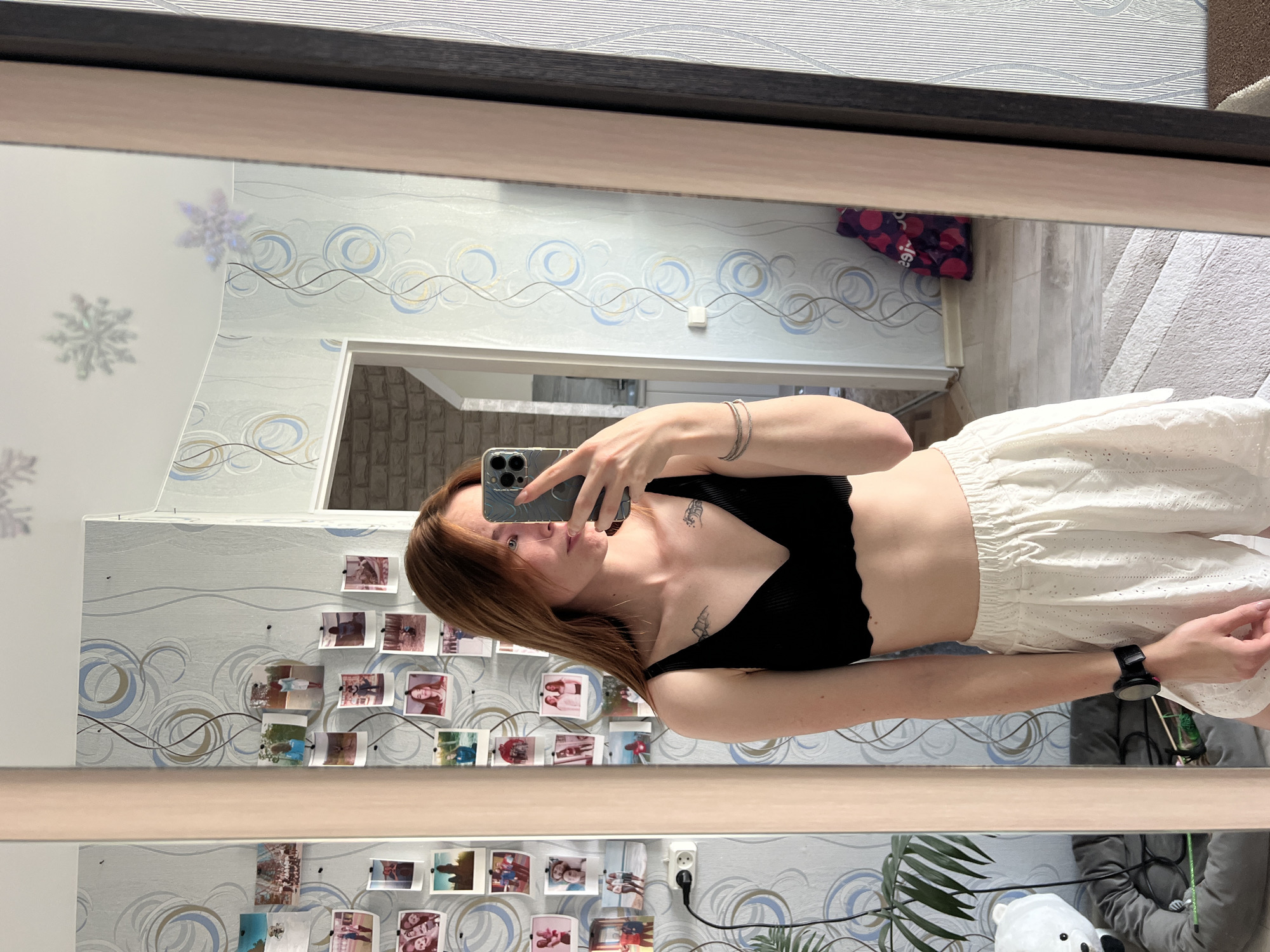 The image shows a woman taking a selfie in a mirror. She is wearing a black top and white shorts. The background includes a wall with patterned wallpaper and a collection of photos. The location is likely Chaikovsky, Russia. Additional details include a doorway, a rug, a power outlet, and a potted plant.
