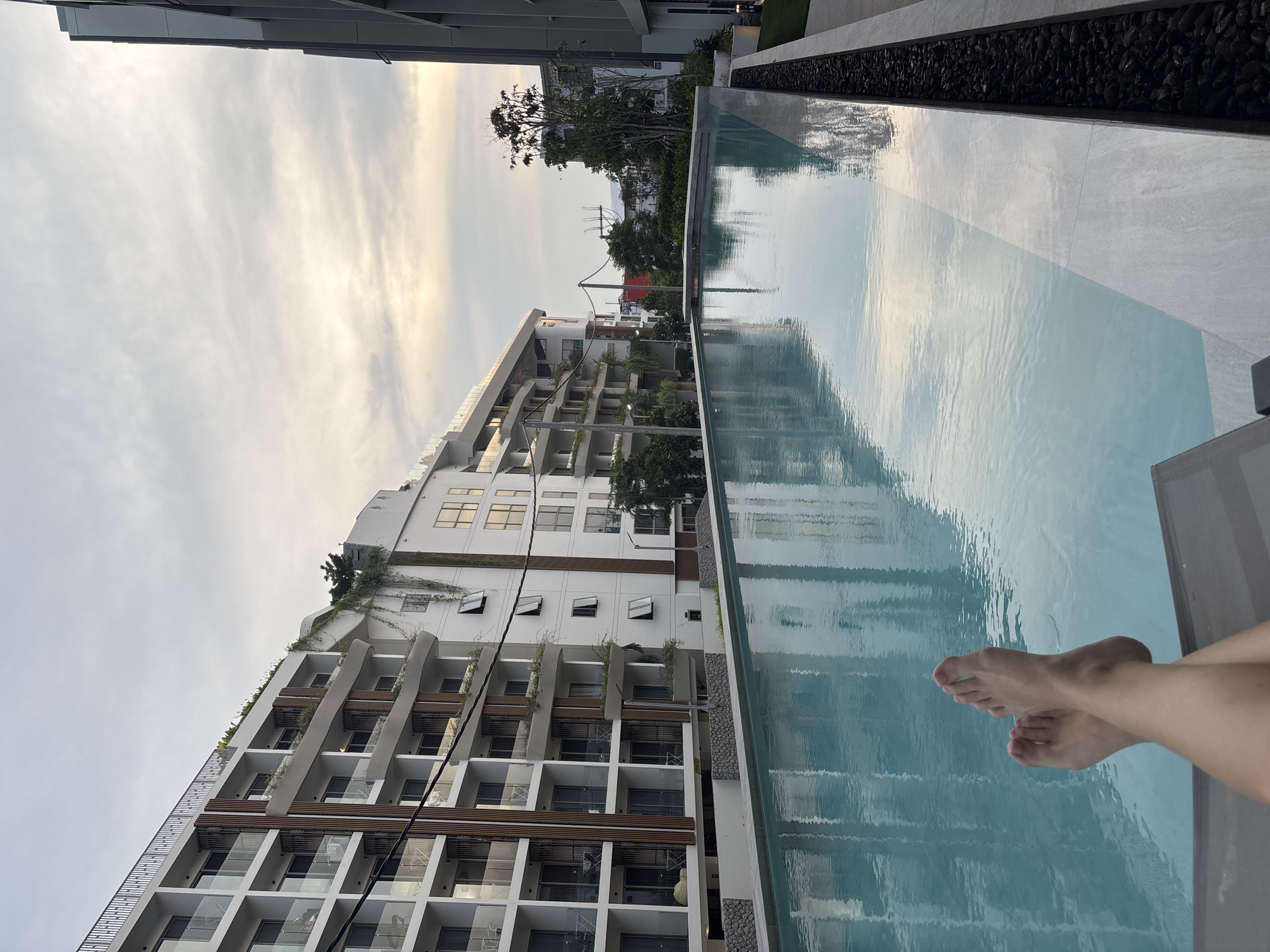 This image depicts a serene poolside scene at what appears to be a modern hotel or resort in Choeng Thale, Thailand, during the late afternoon or early evening, characterized by soft, warm light in the sky.
The primary subject is a pair of human feet, presumably belonging to someone relaxing on a lounge chair (partially visible on the right), with their feet positioned close to, or possibly just in, the tranquil blue water of an infinity-edge swimming pool. The water’s surface reflects the bright, partly cloudy sky and the surrounding structures, creating gentle ripples.
In the background, a large, contemporary multi-story building with a white facade dominates the left side of the frame. It features numerous balconies with glass railings and dark wooden elements, suggesting a modern architectural style. Greenery is visible cascading down parts of the building and on its roof. Beyond this building and in the mid-ground, trees and utility poles with overhead power lines can be seen. The immediate edge of the pool features light-colored tiling and a narrow strip filled with dark, smooth stones. The sky is bright with subtle variations in cloud cover, consistent with a golden hour or dusk setting. There is no visible text in the image.