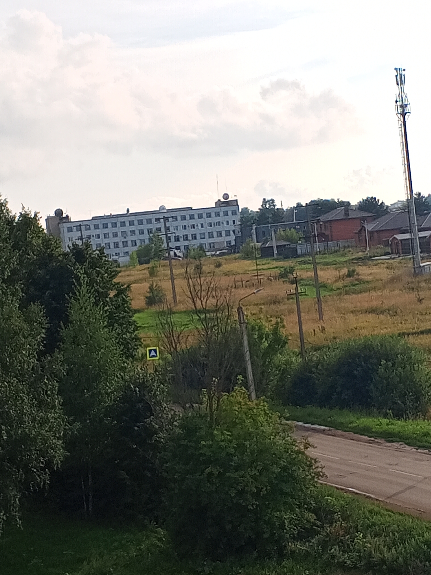 The image depicts an outdoor scene in Chistopol, Russia. A large, multi-story building with many windows dominates the background. In the foreground, there is a road with white lane markings, bordered by lush green vegetation and trees. A yellow triangular sign with a white arrow is visible on the side of the road. A tall cell tower stands prominently on the right side of the image, with a communication tower also visible behind the building. The sky is cloudy, with patches of white and gray. The overall scene appears to be a semi-urban or suburban area, possibly on the outskirts of Chistopol.