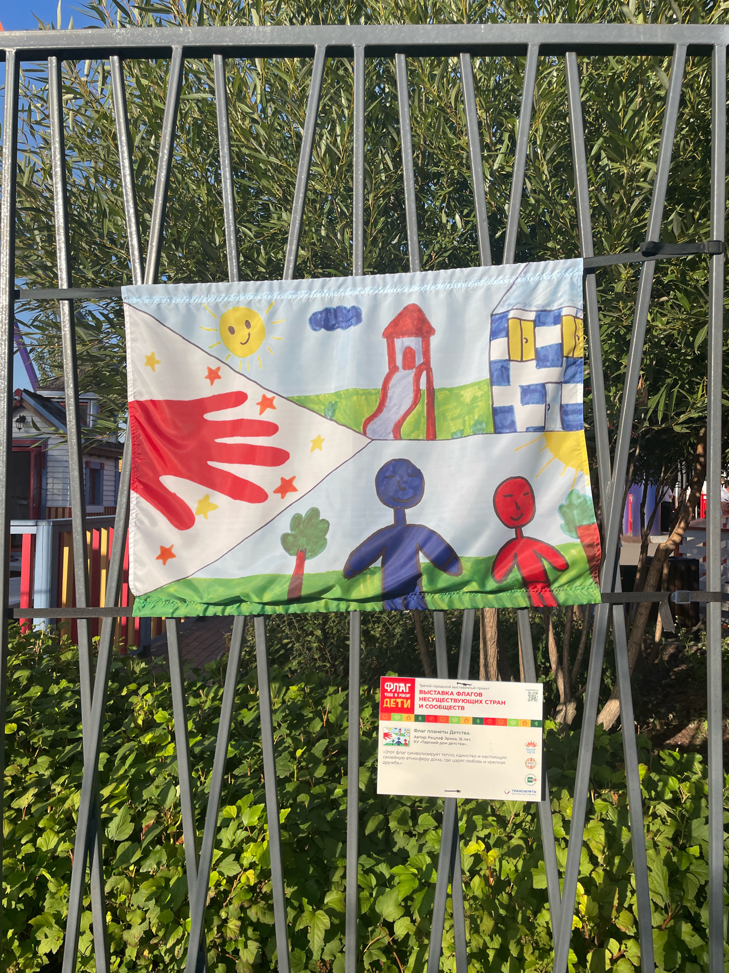 A flag, likely an art project, is displayed on a metal fence with a background of green foliage and a bright blue sky. The flag is divided into three sections. The top left depicts a red handprint with yellow and orange stars and a yellow sun with a smiley face and clouds. The top right section shows a red slide structure and a house with blue and white checkered walls and yellow doors. The bottom section features two stick figures, one blue and one red, standing in a grassy area with a small green tree and another tree on the right. A sign is attached to the bottom right of the flag, providing information in Russian, indicating that this is part of an exhibition called "Flags of Non-Existent Countries" by children. The text on the sign mentions the project name "Flags of Children," the exhibition title "EXHIBITION OF FLAGS OF NON-EXISTENT COUNTRIES AND COMMUNITIES," the author "Author: Reuylay Irinia, 16 years old," and the institution "KU 'Tatarsky Dom Detstva'". The exhibition appears to be a city-wide project. The scene is set outdoors, with the fence and foliage suggesting a public space such as a park or school grounds in the city of Omsk, Russia.