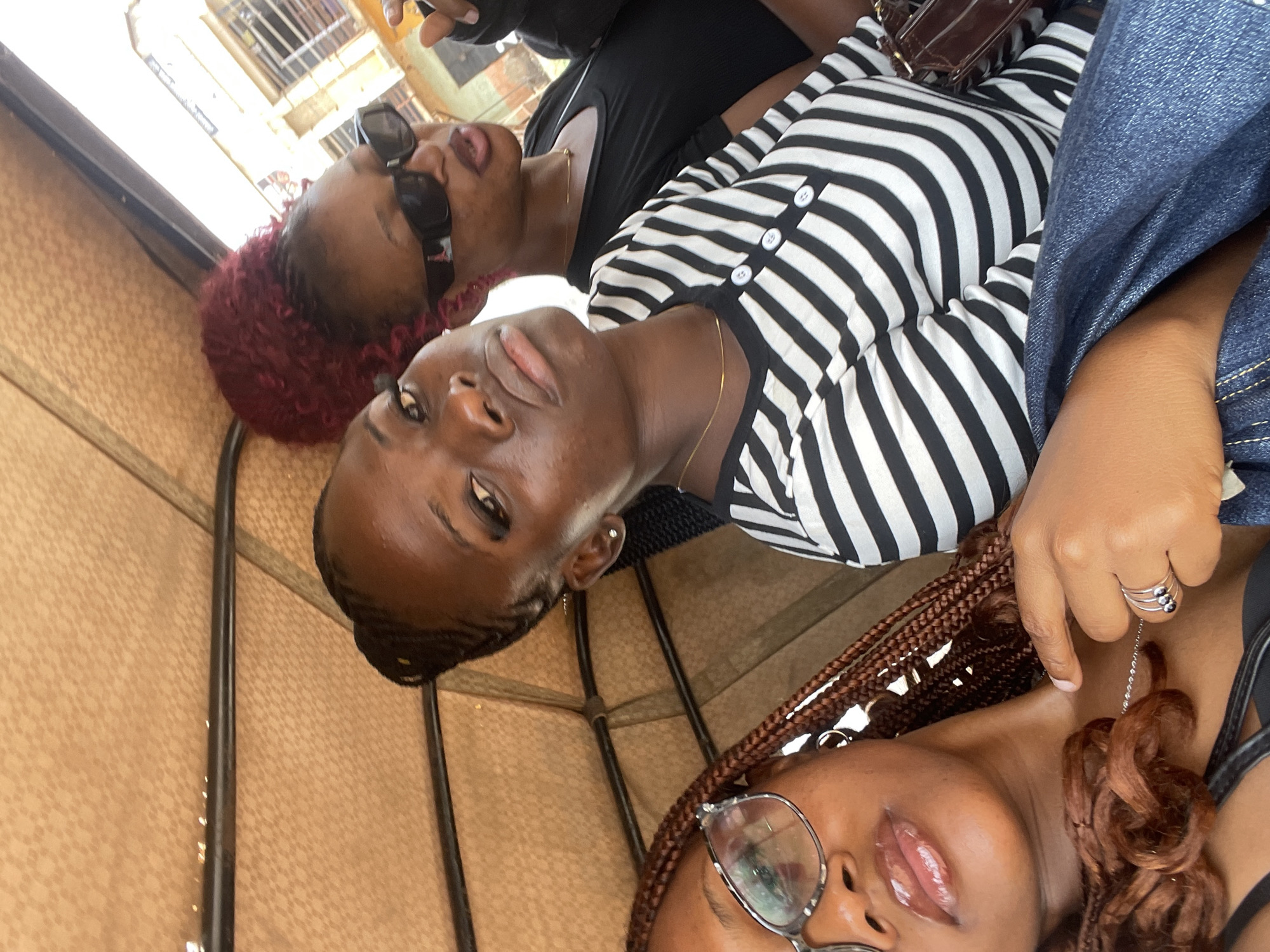 Three young Black women are seated in the back of a motorized rickshaw, a common mode of transport in many Nigerian cities, including Rafin Par. The rickshaw's interior is lined with a patterned tan fabric, and a black metal bar runs across the top.
The woman in the foreground, closest to the camera, is wearing glasses with a silver frame and has her hair braided in long, dark brown plaits. She is looking directly at the camera with her lips slightly parted and glossy. She is wearing a black top and a blue denim jacket draped over her shoulders, and a silver necklace with a pendant.
The woman in the middle is wearing a black and white striped shirt with three white buttons at the collar. She has dark hair braided away from her face and is looking to her right. She is wearing a thin gold necklace and a small earring in her left ear.
The woman in the background, to the right, has bright red curly hair styled in a bun. She is wearing black sunglasses and a black top. She is looking forward and appears to be speaking. Her left arm is visible, and she is wearing a ring on her left ring finger.
The background visible through the open side of the rickshaw shows buildings with signs and windows, suggesting an urban environment in Rafin Par. The overall lighting suggests it is daytime. The scene is candid, capturing a moment of shared travel.