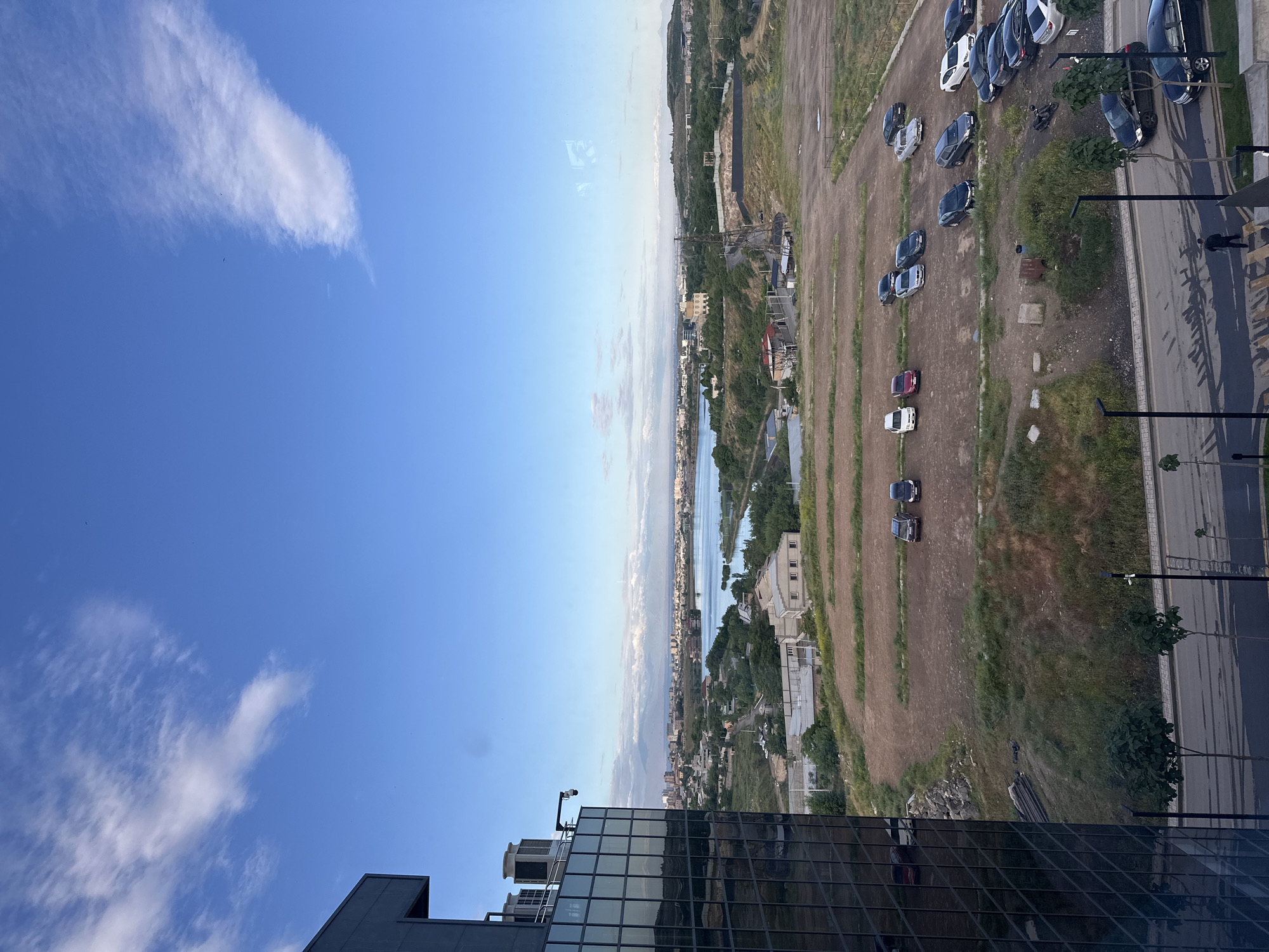 The image shows a view from a high building in Yerevan, Armenia. In the foreground is the top of a modern building with glass windows, likely part of the building from which the photo was taken. Beyond the building is a mostly vacant lot, with rows of parked cars. Further in the distance is a river running through the city, with buildings and trees along its banks. The sky is blue with scattered clouds.
