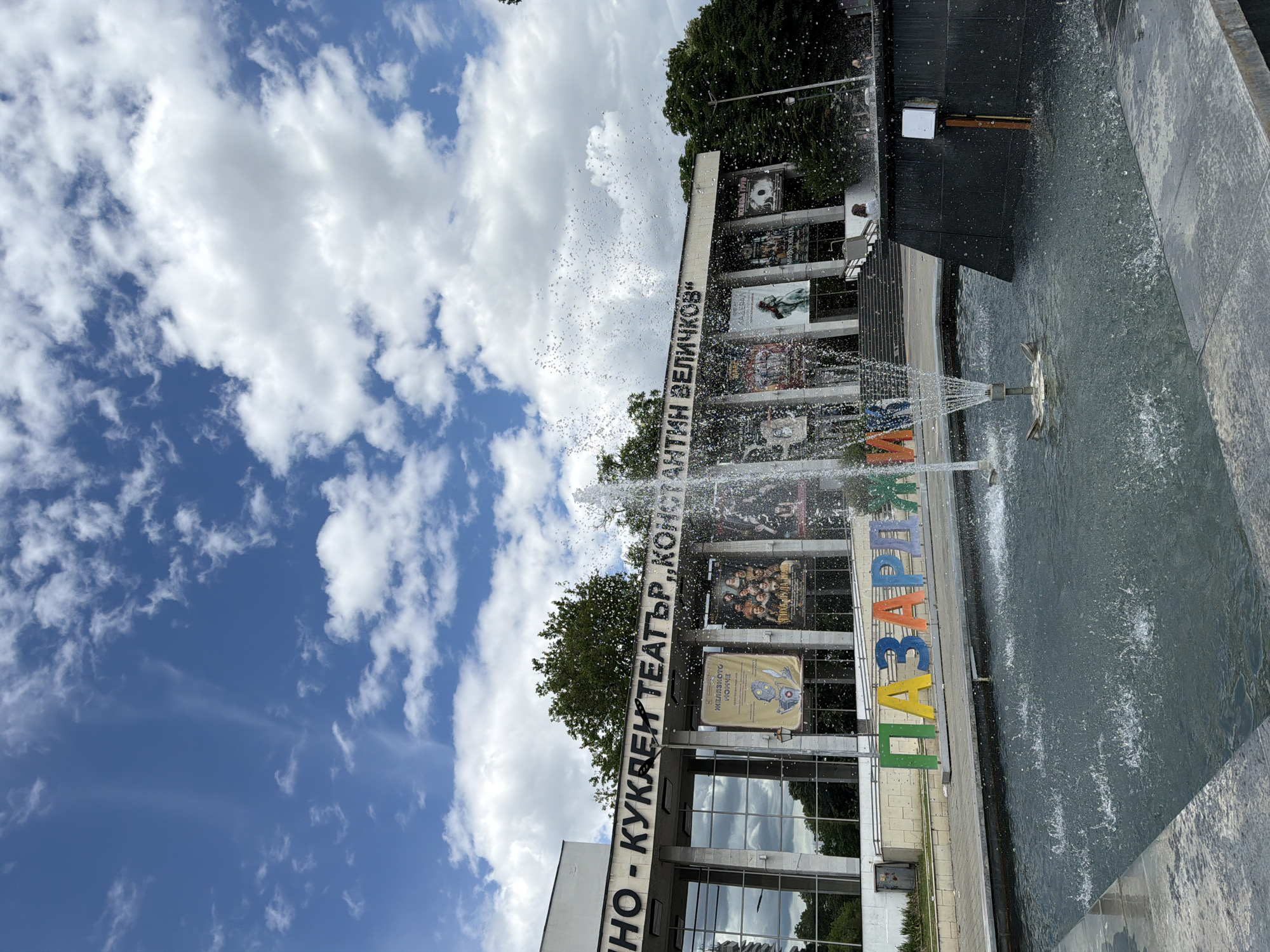 The image captures a view of the "Konstantin Velichkov" Drama Theater in Pazardzhik, Bulgaria, on a cloudy day. In the foreground, a fountain with multiple spray jets is active, casting water droplets into the air. In front of the theater, large, colorful, three-dimensional letters spell out "PAZARDZHIK". The theater itself has a modern facade with large glass windows and a marquee displaying posters for theatrical productions. Several trees are visible behind and to the sides of the building. The sky is filled with scattered white clouds against a blue background. A few people are present in the background, near the theater entrance and a black structure to the right of the fountain.