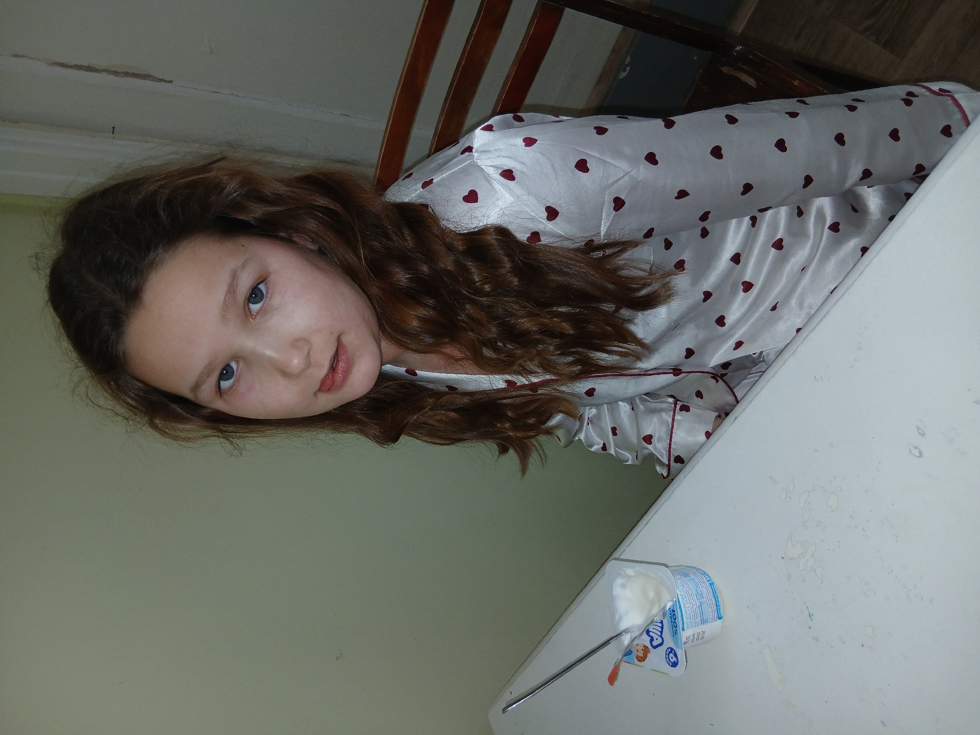 A young girl with long, wavy, reddish-brown hair and blue eyes looks directly at the camera with a slight, gentle smile. She is wearing white satin pajamas patterned with small red hearts and red trim. The image is taken from a slightly tilted perspective, with her head leaning towards the lower right.
Behind the girl, part of a wooden chair back is visible. To the left of her, a plain, light-colored wall forms the background. In the foreground, on the right side of the image, is a white tabletop with some faint smudges or spills. On the table, an open plastic container of yogurt with a spoon in it is visible. The container is white and blue, and features text including "100%" and "АГУША" (Agusha) in Cyrillic, along with a number "5" in a circle. The scene appears to be an indoor, casual setting, possibly a home, capturing a quiet moment, perhaps during a snack. The lighting is artificial, offering no specific cues regarding the time of day.