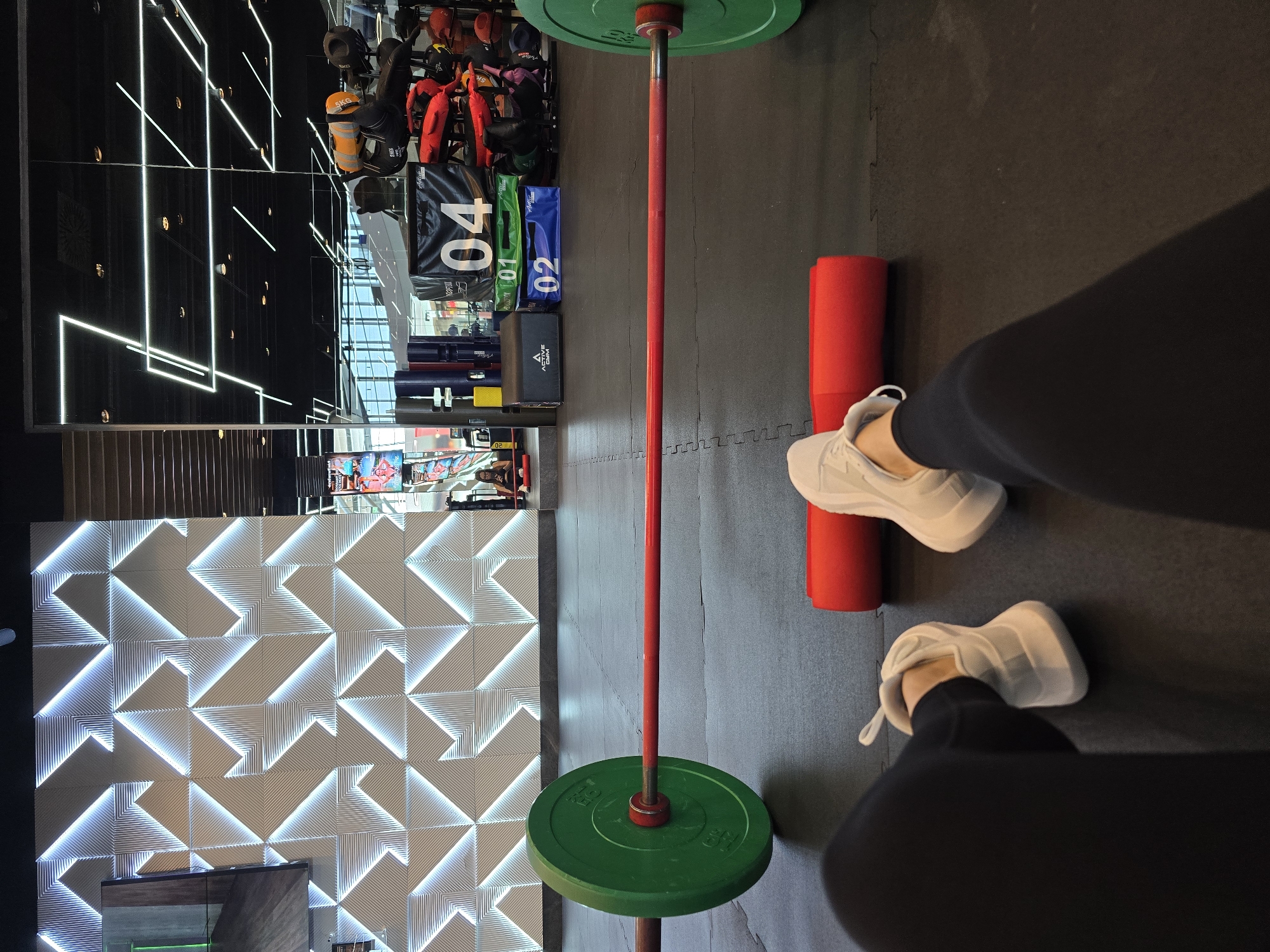 A first-person perspective photograph shows a person standing in a gym setting with a barbell positioned centrally in the frame. The barbell has green weight plates on both ends and a red-painted bar. A red foam roller lies on the floor near the person's feet. The person is wearing black leggings and white sneakers.
The background reveals a gym interior. A wall with a geometric pattern of illuminated triangles is visible to the left. Mirrored panels reflecting the gym's ceiling and various equipment, including weight bags and training aids, are also visible. The scene suggests a well-equipped fitness facility.
It's not possible to confirm a location in Burgas, Bulgaria from the image.