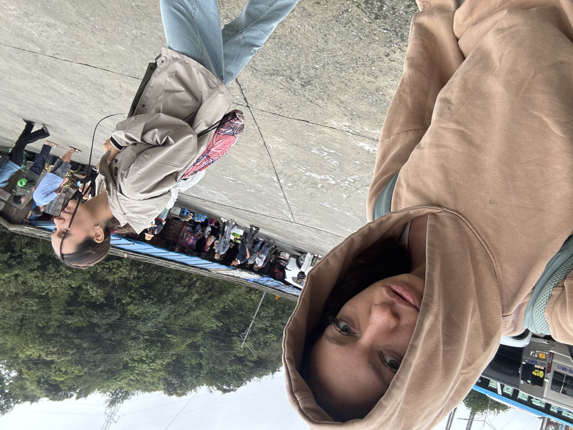 The image appears to be a selfie taken by a woman wearing a tan hoodie. In the background, another woman is visible wearing jeans and a light-colored jacket with a pink bag. Both women appear to be on a paved surface possibly a sidewalk or street. Behind them is a busy street scene with people, stalls, and buses, partially obscured by green foliage. The location appears to be in Binh Lu, Vietnam, based on the writing on a bus in the background.