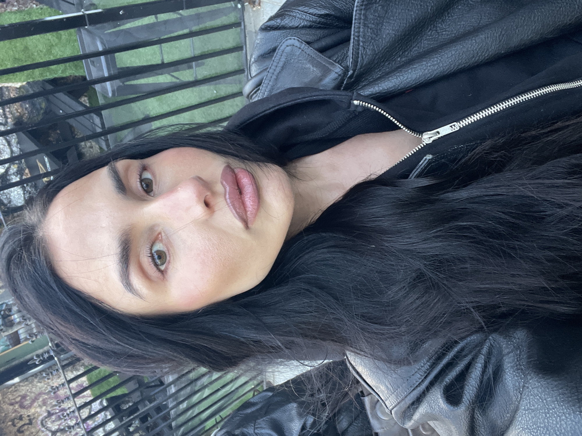 A close-up, rotated portrait image features a young woman with long, dark, wavy hair cascading over her shoulders. She has light skin, prominent dark eyebrows, and light brown or hazel eyes looking directly towards the viewer. Her lips are full, appearing to have a neutral-toned lip gloss or lipstick applied. A subtle blush is visible on her cheeks. Her expression is calm and direct.
She is wearing a black leather jacket with a textured surface, layered over a black garment, possibly a hooded sweatshirt, with a silver zipper clearly visible on the front. Her right shoulder and part of her arm are visible within the frame.
The background, somewhat out of focus, suggests an outdoor setting, consistent with London, United Kingdom. A black metal railing or fence spans horizontally across the upper portion of the frame (when viewed in its original upright orientation). Beyond the railing, patches of green grass or foliage are visible, along with indistinct shapes that could be urban landscaping or outdoor furniture. The lighting appears to be natural daylight, indicating it is daytime, possibly a soft, overcast day given the lack of harsh shadows. No visible text is present in the image.