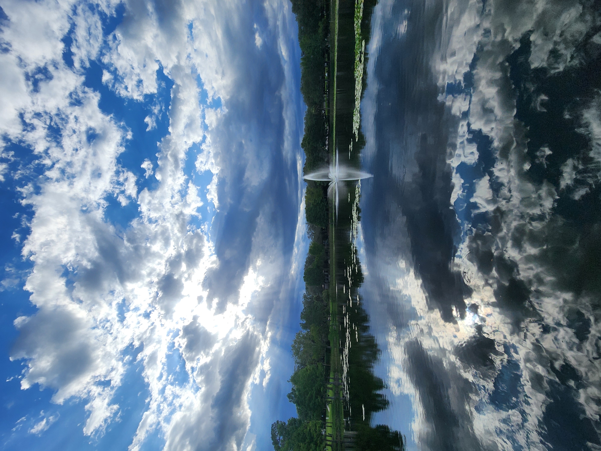 The image depicts a serene lake scene with a fountain in the distance, located in the city of Maitland, United States of America. The sky is filled with dramatic, dynamic clouds, with sunlight piercing through in rays, creating a play of light and shadow. The lake's surface acts as a mirror, reflecting the sky, clouds, and the surrounding treeline with remarkable clarity. Scattered lily pads are visible on the water's surface. The treeline consists of lush green foliage, suggesting a park or natural area within the city. The overall atmosphere is peaceful and picturesque, with a sense of natural beauty. No people are visible in the image.