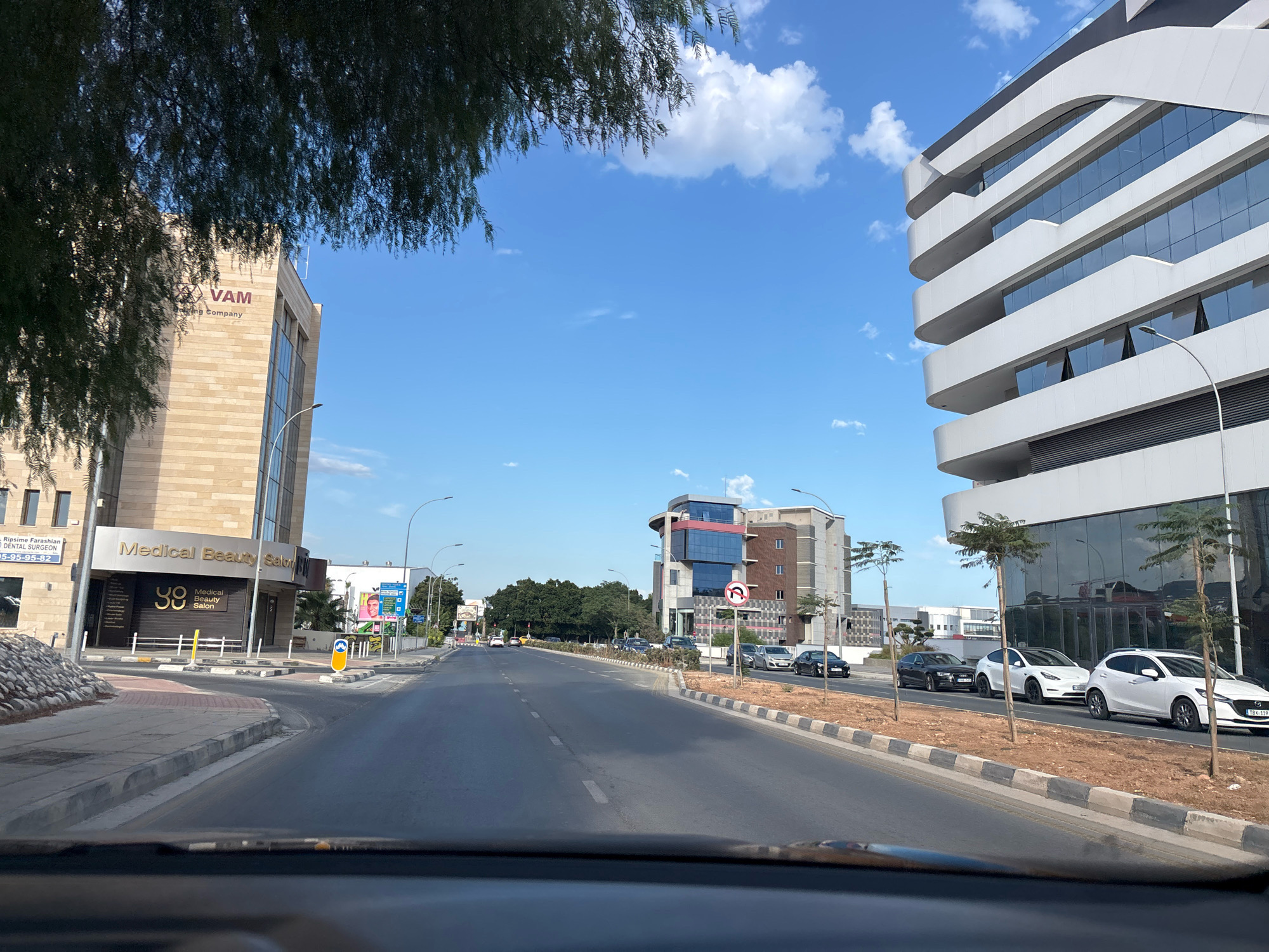 The image depicts a street scene in Mesa Geitonia, Cyprus, on a clear, sunny day. The perspective is from within a vehicle, looking forward. On the left side of the road, a light-colored, modern building with the signage "VAM" and "Medical Beauty Salon" is visible. Adjacent to this building, a low, stone-covered embankment lines the sidewalk. Across the street, a white, multi-story building with a distinctive curved facade and large glass windows dominates the right side of the frame. The road is asphalt, with white lane markings and a dedicated turning lane marked by bollards. Several cars are present on the road and parked along the sidewalk, including a white Tesla and a white Mazda. Trees are planted along the median and sidewalks. In the background, other buildings and more trees can be seen, with a bright blue sky with scattered white clouds overhead. Traffic signs, including a no-U-turn sign, are visible. The overall scene is one of urban development and daily life.
