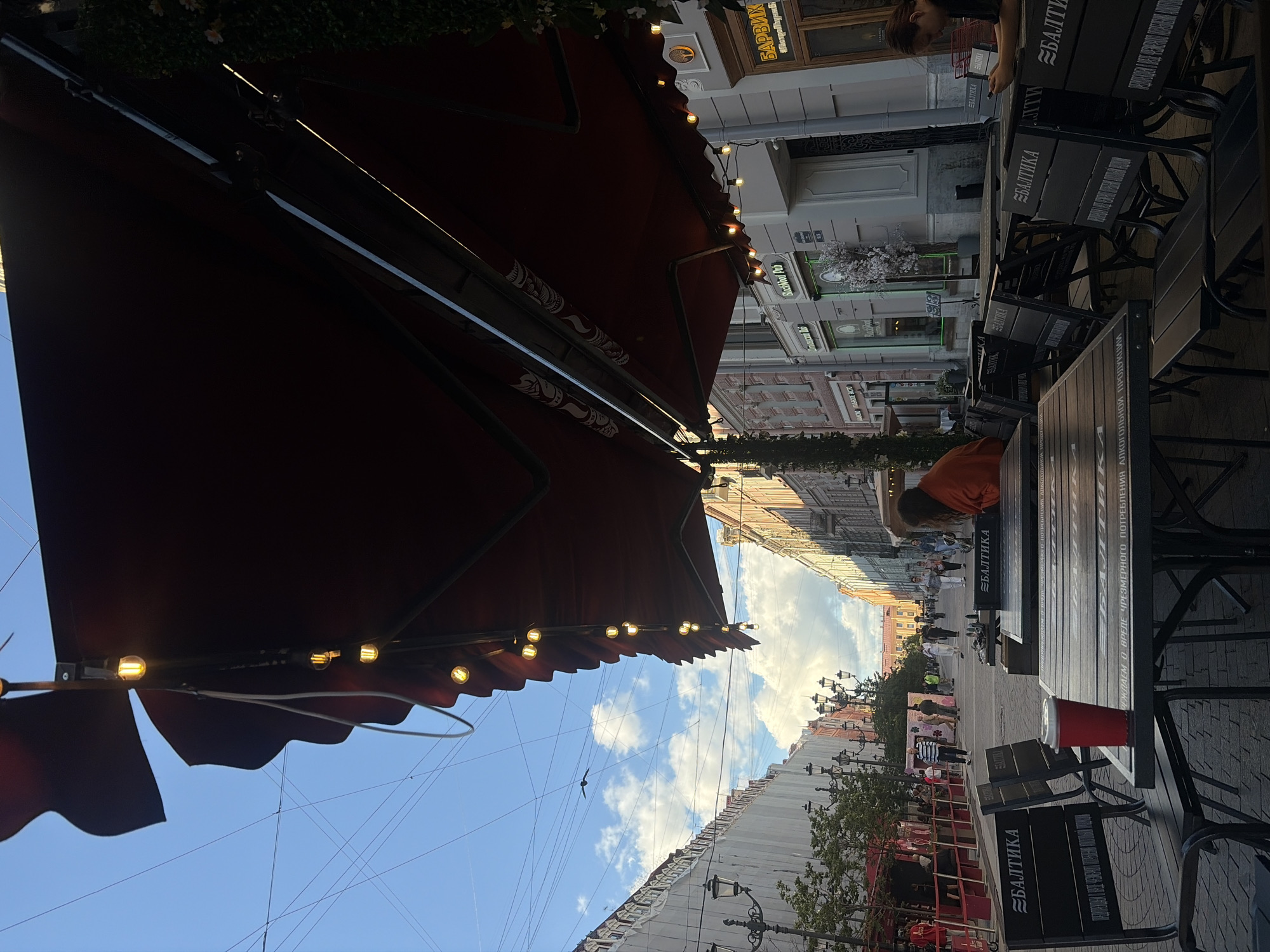 The image captures an outdoor seating area of a establishment, likely a cafe or restaurant, on a street in Saint Petersburg, Russia. The perspective is from under a large, dark red awning, adorned with string lights that are illuminated. The awning creates a shaded foreground, contrasting with the bright sky and clouds visible above.
In the midground, a street scene unfolds. Several tables and chairs, some with benches, are set up. The tables appear to be made of wood with metal frames, and some are branded with the Cyrillic text "БАЛТИКА". On one table, a red paper cup with a white lid is prominently placed. A person in an orange top is seated at one of the tables, looking away from the camera. Further down the street, other patrons are seen walking and sitting.
The architecture of the buildings lining the street suggests a European urban setting, with multi-story buildings featuring classical architectural elements. Signs in Russian are visible on the buildings, including "БАРВИХ" which indicates a location or establishment. The presence of street lamps and trees adds to the urban ambiance.
The overall mood seems relaxed and casual, with people enjoying an outdoor setting. The time of day appears to be late afternoon or early evening, given the lighting conditions and the illuminated string lights. The sky is a clear blue with scattered white clouds. The weather seems pleasant for an outdoor dining experience. The visible text on the tables and signs points to a Russian context, consistent with the specified location of Saint Petersburg.
