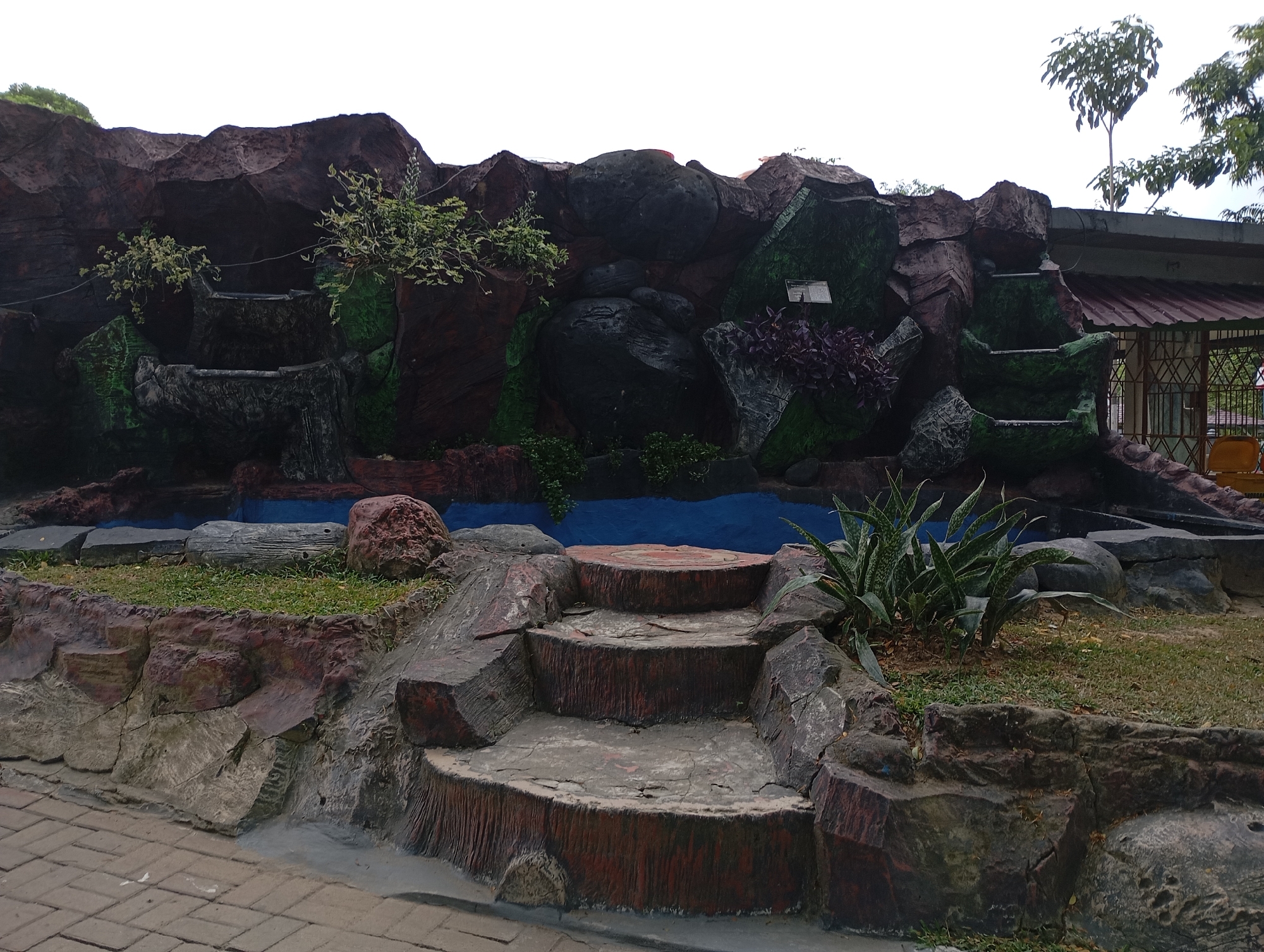 The image depicts a man-made rock formation with a small waterfall or tiered water feature in a park-like setting. The rocks are textured and painted in various shades of brown, red, and green, simulating a natural landscape. There are several tiers of "waterfalls," but no actual water is visible. In front of the rock formation, there are three steps made to look like logs, leading up to a blue pool area. Grass and several plants, including snake plants, are growing around the rocks and on top of some sections. The ground in front of the steps is paved with brick-like stones. In the background, a building with a reddish roof and a fence can be seen. The sky is overcast. This scene is likely located at the Balikpapan Ocean Square, a public park in Balikpapan, Indonesia, known for its artistic rock installations and recreational spaces.