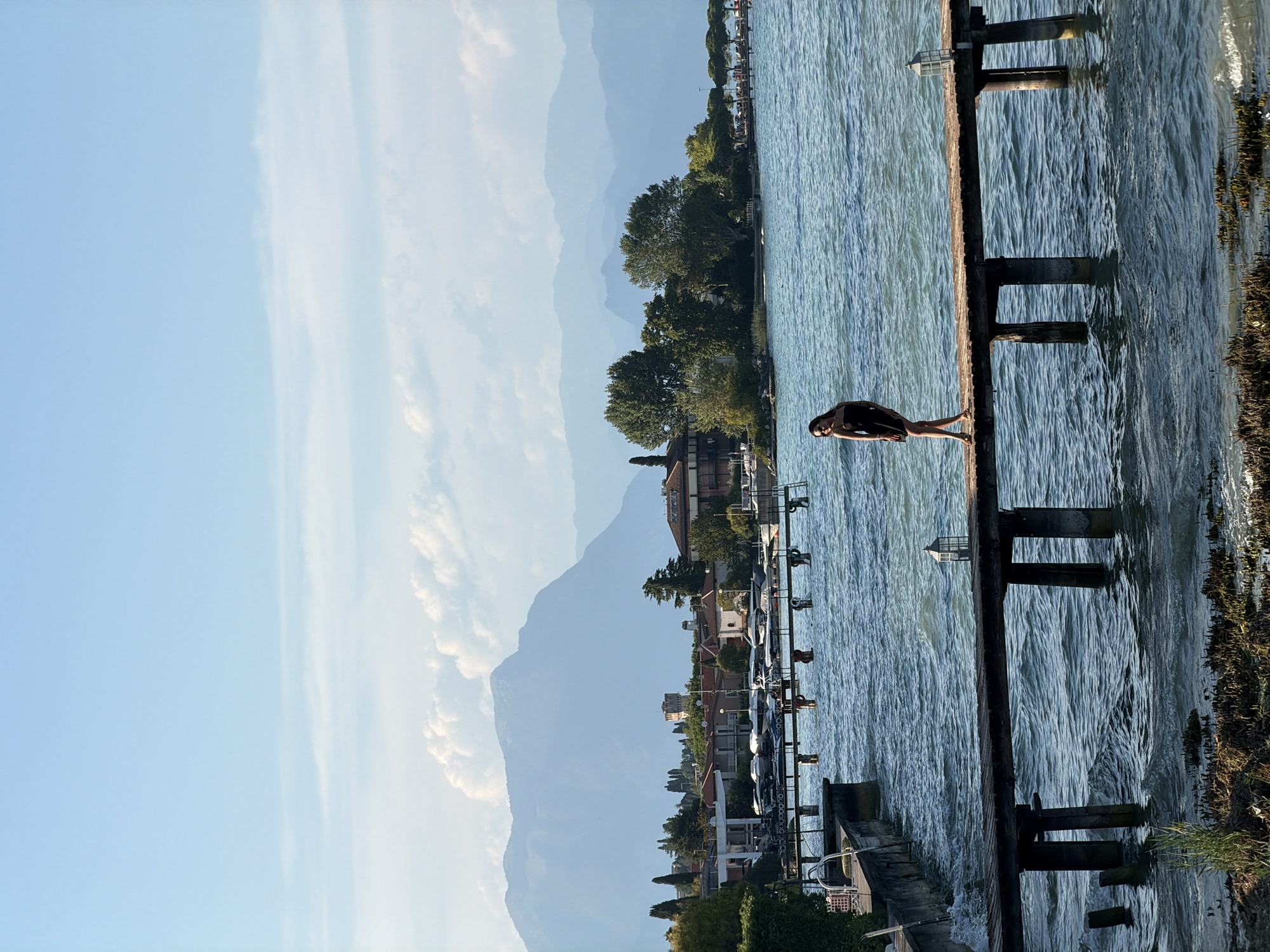 A full-shot vertical photograph captures a woman in a black dress standing on a wooden pier extending into a body of water. The woman faces left, looking towards the viewer with sunglasses perched on her head and her left hand on her hip. The pier is supported by dark, weathered wooden pilings that disappear into the choppy blue water. Further back, a second, longer pier with multiple pilings is visible, and beyond that, a shoreline with buildings, trees, and a harbor filled with boats. In the distance, layered mountain ranges fade into a pale blue haze under a bright, clear sky with scattered white clouds. The scene is set during daylight hours, suggesting a mild climate.