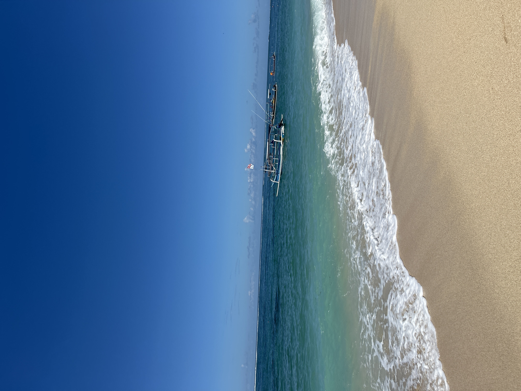 The image shows a beach scene in Kuta Selatan, Indonesia. A clear blue sky occupies the left side of the frame. The ocean extends from the middle to the right, transitioning in color from deep blue to turquoise closer to the shore. White waves are breaking along the sandy beach, which occupies the rightmost section of the image. Several outrigger boats are visible in the water, slightly off-center. One boat has a small Indonesian flag.