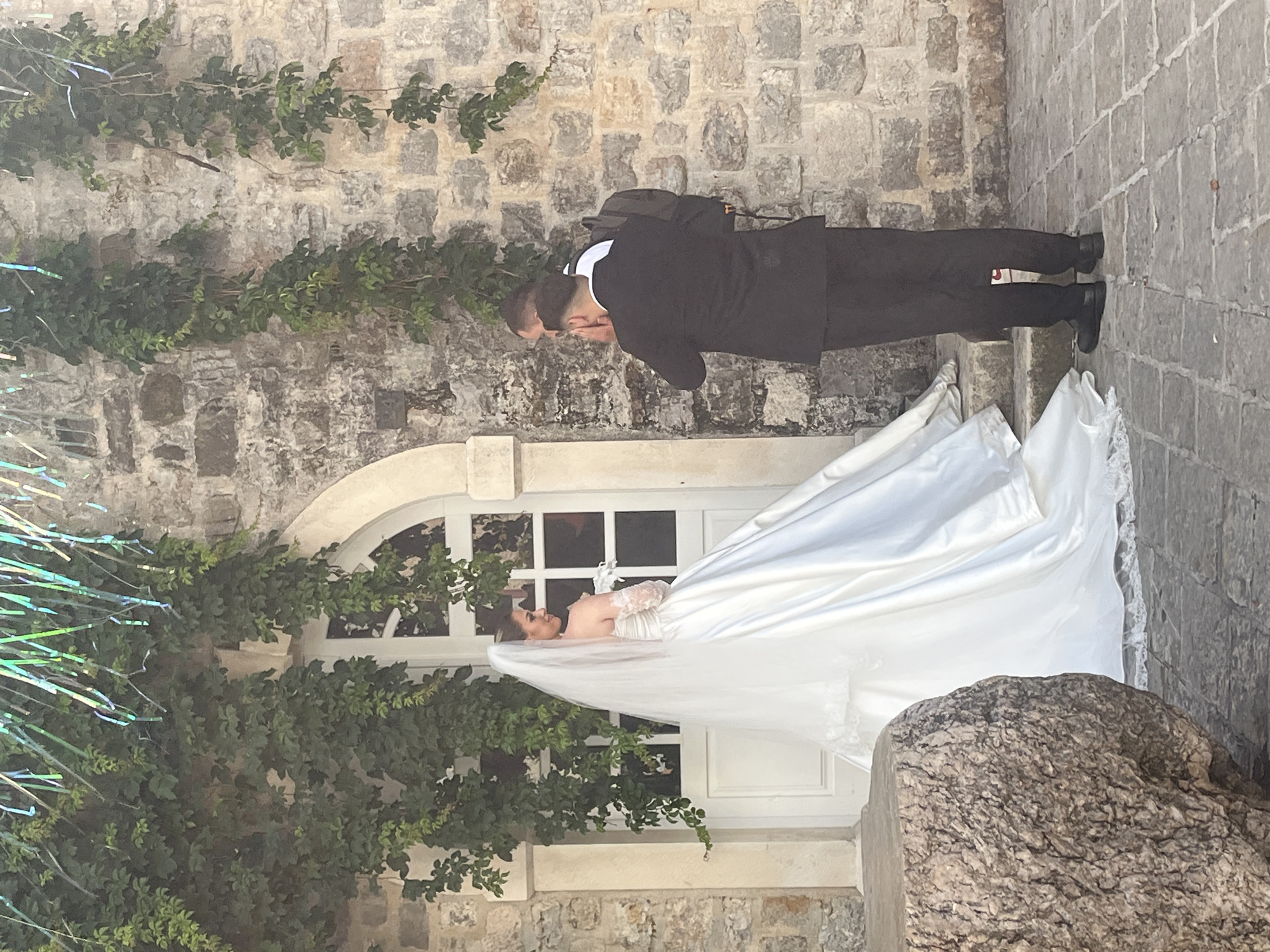 This full-shot image, taken in Budva, Montenegro, depicts a bride and groom outdoors, likely during a wedding event or photoshoot. The scene is set against a textured stone wall, characteristic of an old town or historic area, with green ivy and vines growing across it.
On the left side of the frame, the groom stands, mostly facing away from the camera. He is dressed in a black suit, a white shirt, and dark shoes, and wears a black backpack. His hands are raised towards his face or neck, suggesting he might be adjusting something or having a moment of reflection.
To the right, the bride stands gracefully near a white-framed window, looking upwards and slightly to her right with a serene expression. She is adorned in a beautiful white wedding gown featuring lace sleeves and a long, flowing white veil that billows around her. The dress and veil are prominent, creating a sense of movement.
The background features the rugged stone wall, an arched white window frame behind the bride, and a paved stone ground underfoot. In the lower right foreground, a large, rough-textured stone is visible. To the far left, faint iridescent streamers or tinsel add a decorative element. The lighting indicates it is daytime. The overall atmosphere is romantic and tranquil, emphasizing the couple within a picturesque, historical setting. There is no visible text in the image.